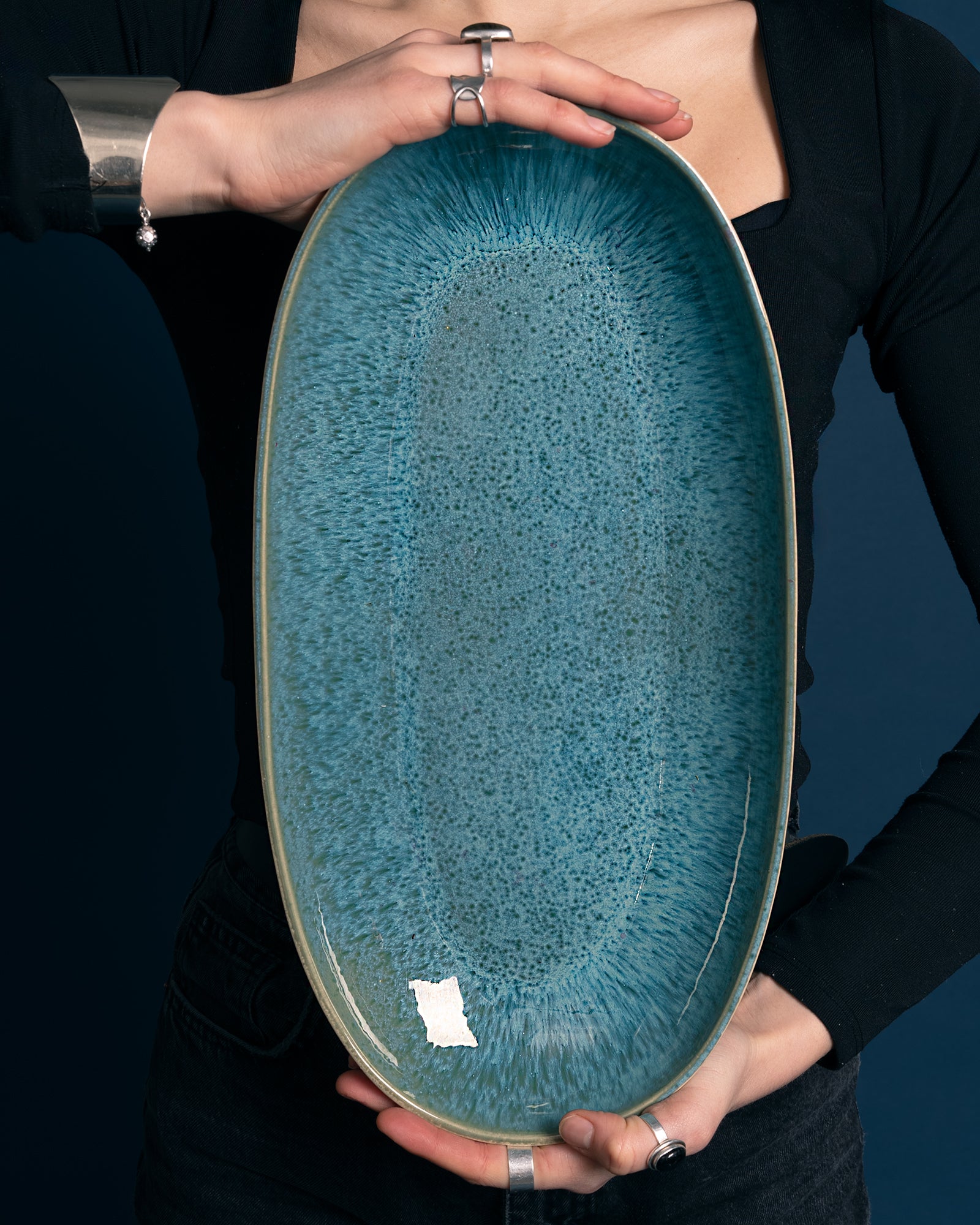 Person hält ein großes, ovales, blau-marmorglasiertes Keramiktablett vor einem dunklen Hintergrund.