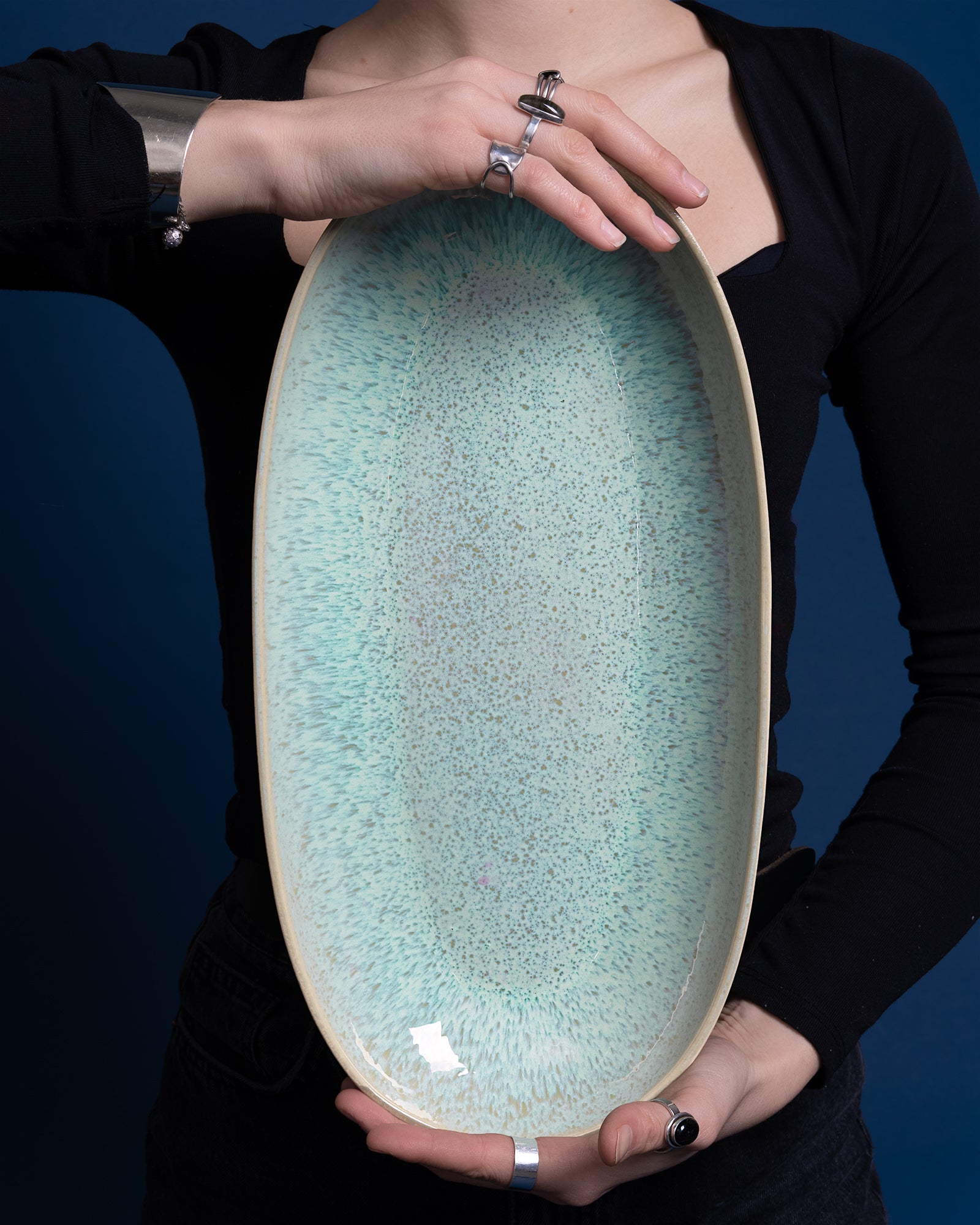 Person hält eine große, ovale, türkis gesprenkelte Keramikschale vor ihrem Oberkörper vor einem blauen Hintergrund.