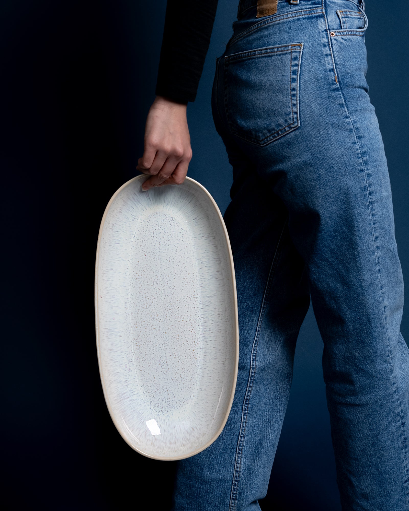 Person in blauer Jeans hält ein großes, ovales, weißes Tablett vor einem dunklen Hintergrund.