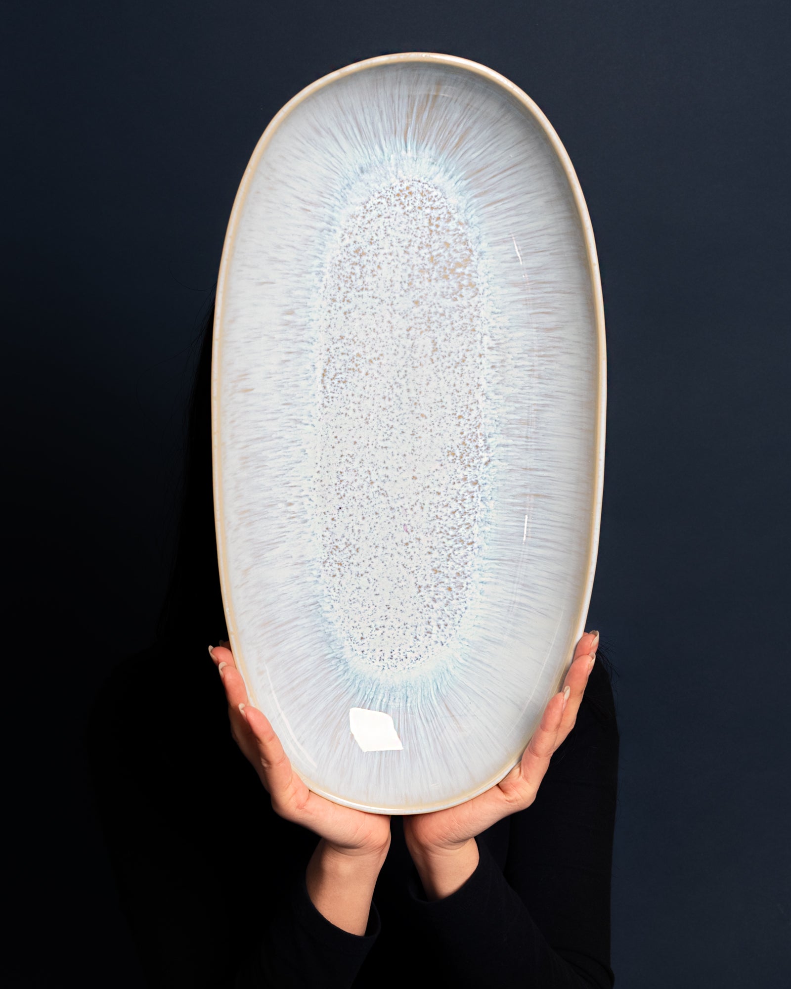 Eine Person hält ein großes, ovales, kunstvoll glasiertes Keramiktablett vor einem dunklen Hintergrund hoch.