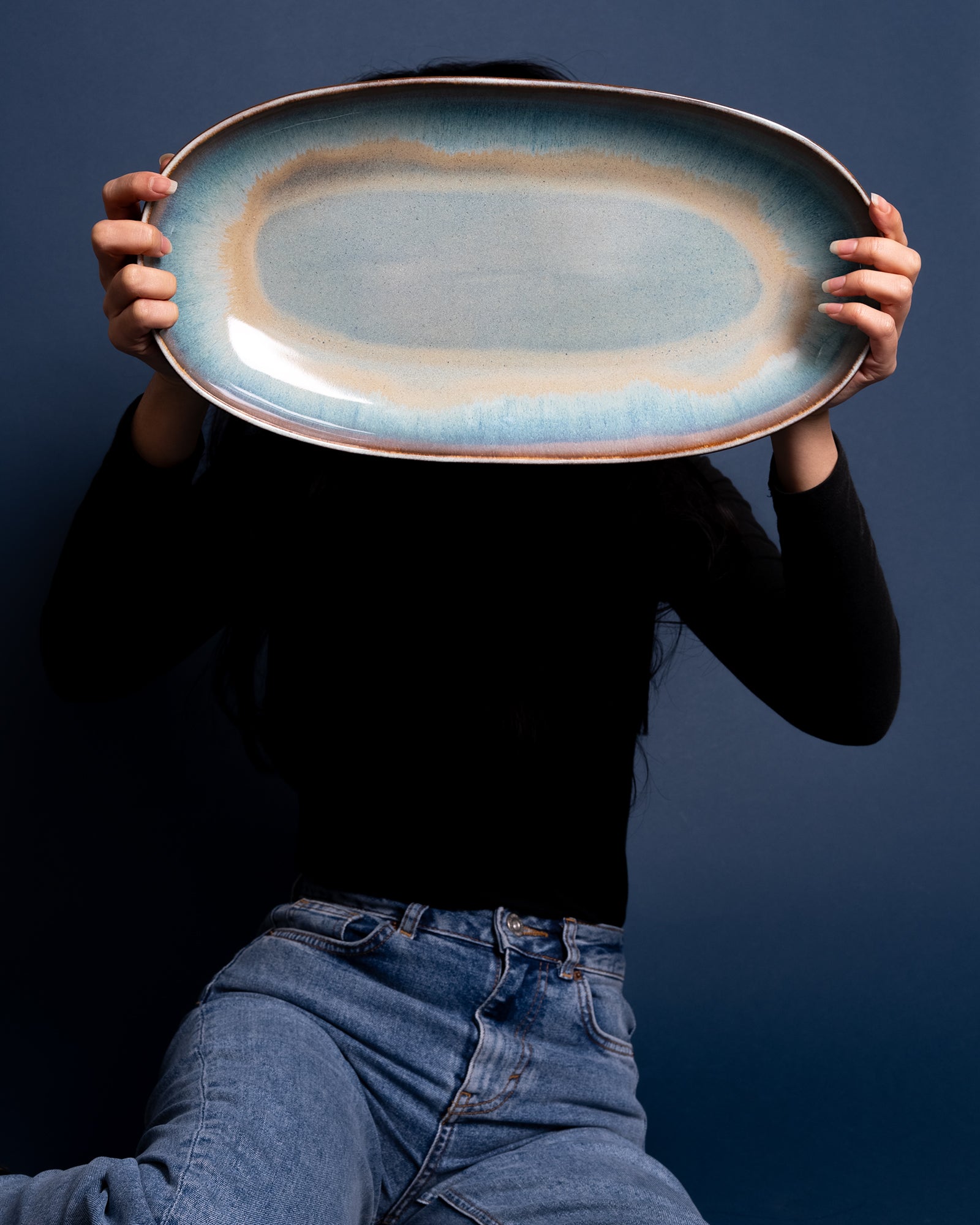 Person in schwarzem Oberteil und Jeans hält einen großen, blaugeränderten Teller vor ihr Gesicht.