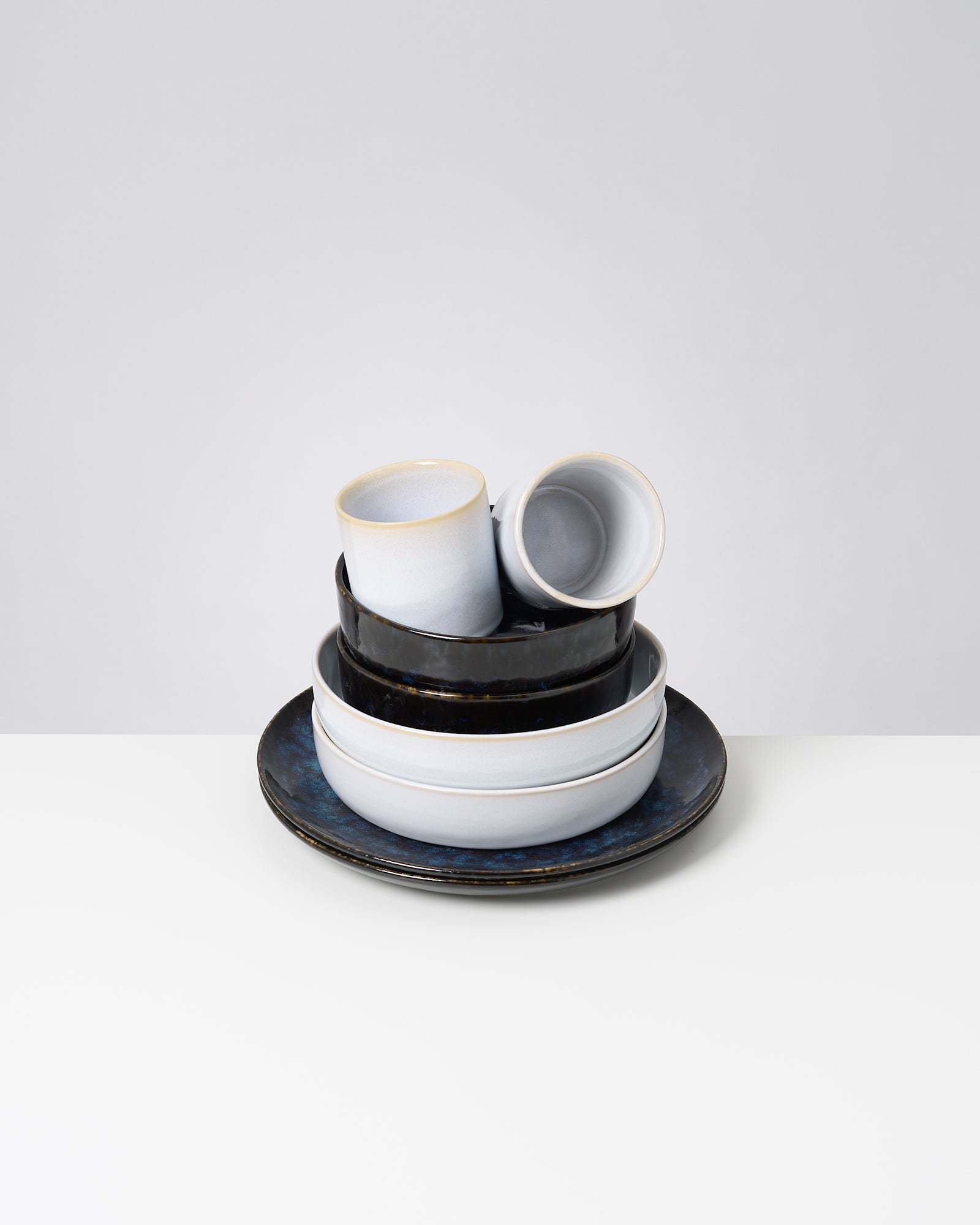 Stapel aus Keramikgeschirr in Blau- und Schwarztönen auf einem weißen Tisch vor grauem Hintergrund.