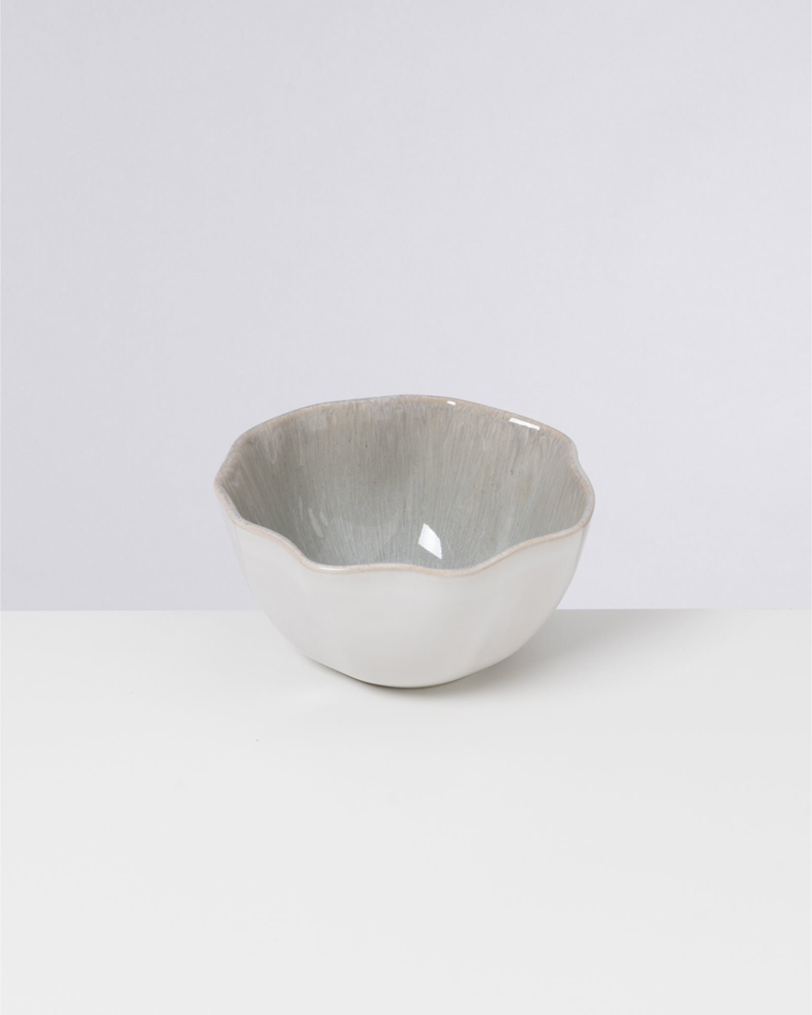 Eine weiße, gewellte Keramikschale steht auf einem weißen Untergrund vor einem grauen Hintergrund.