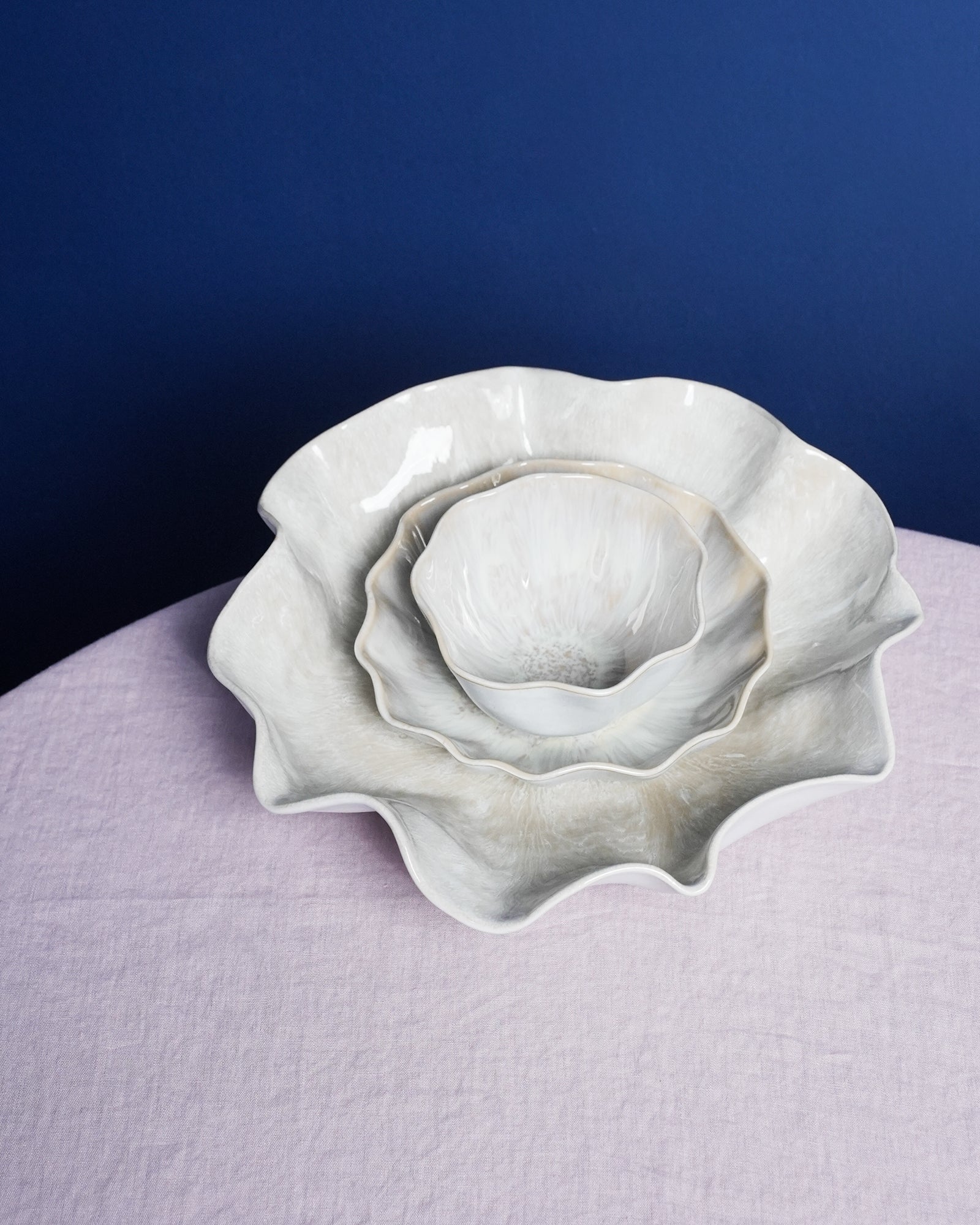 Drei miteinander verschachtelte, blütenförmige Schalen aus Keramik auf einem blasslila Tischtuch vor einem dunkelblauen Hintergrund.