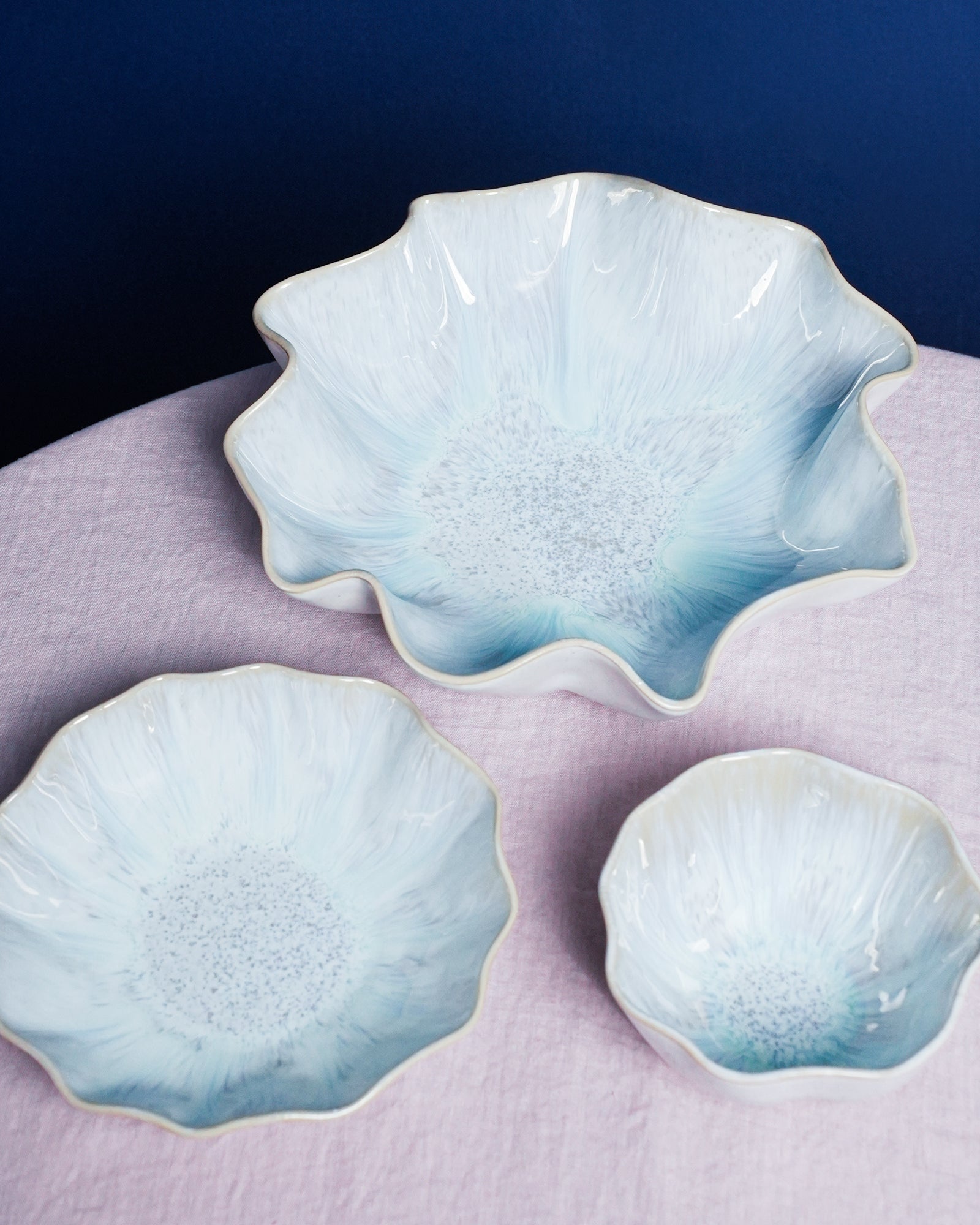 Drei blumenförmige, hellblaue Schalen auf einem lilafarbenen Tisch vor einem dunkelblauen Hintergrund.