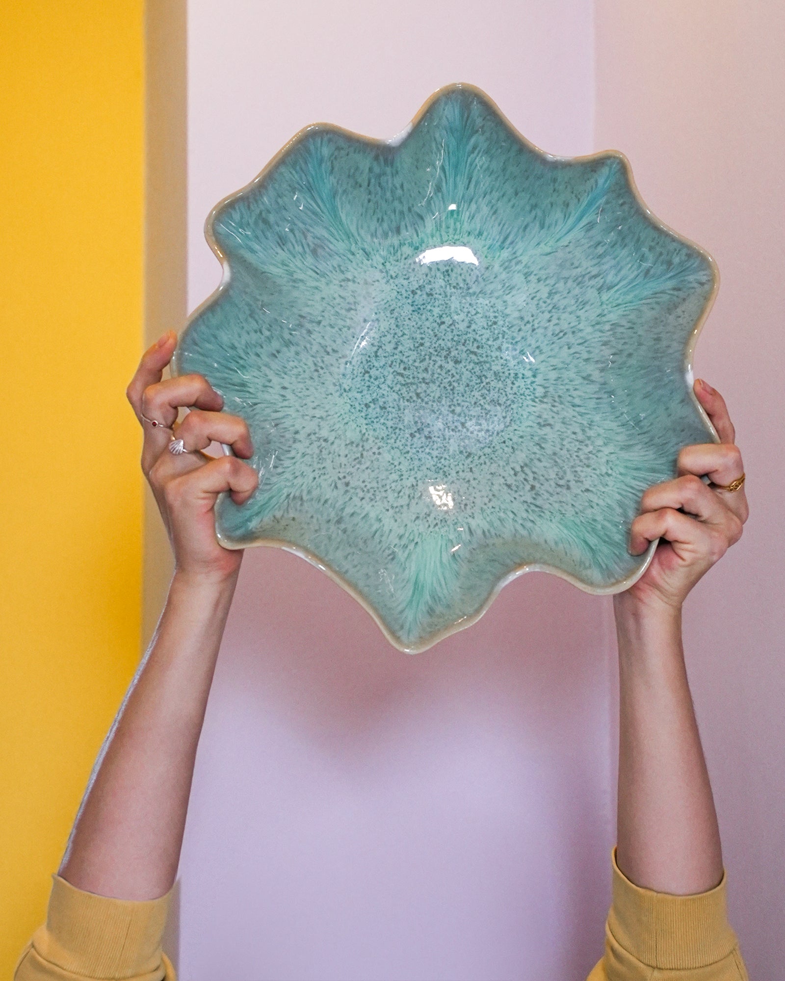 Zwei Hände halten eine große, grüne, sternförmige Schale vor einer zweifarbigen, gelb-rosa Wand.