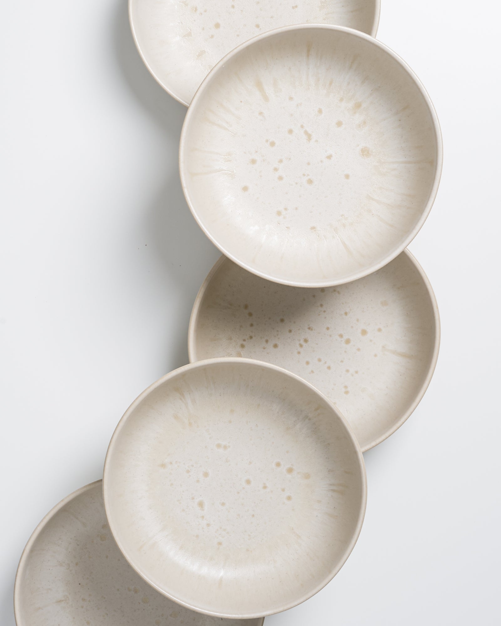 Fünf weiße, gesprenkelte Keramikteller sind in einer diagonalen Linie angeordnet auf weißem Hintergrund.