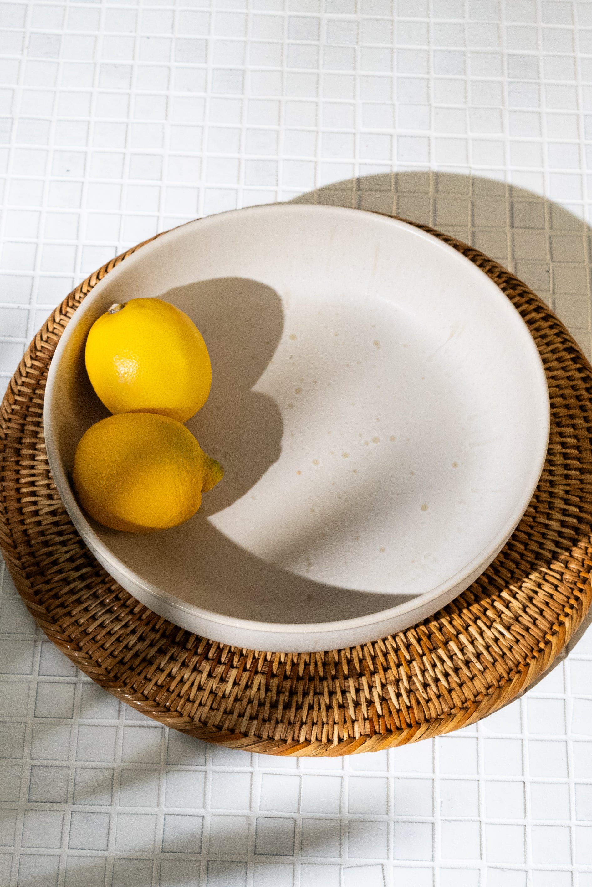 Zwei Zitronen in einer weißen Schale auf einem geflochtenen Untersetzer auf einem weißen Fliesentisch.