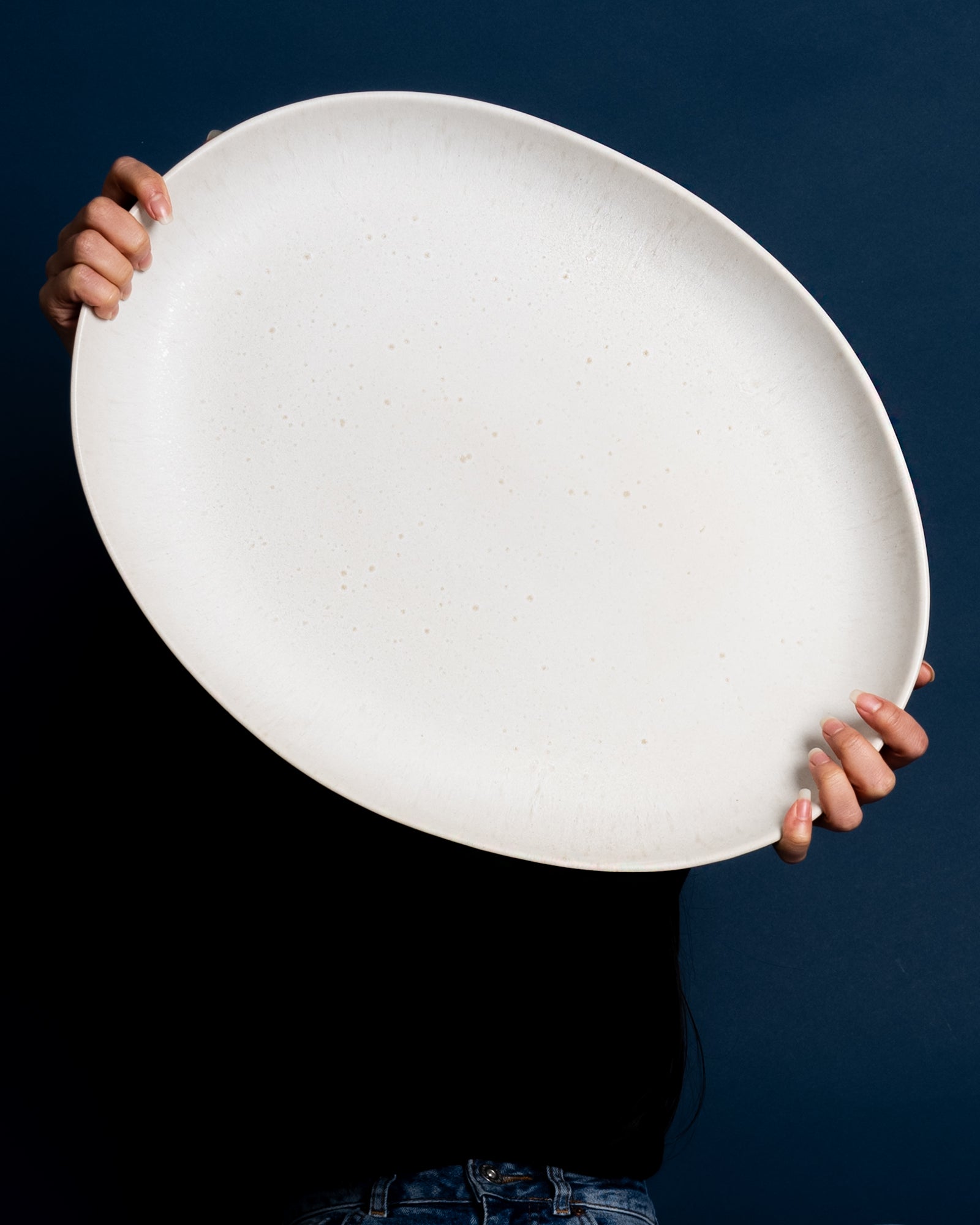 Eine Person hält einen großen, ovalen, weißen Teller vor einem dunkelblauen Hintergrund.