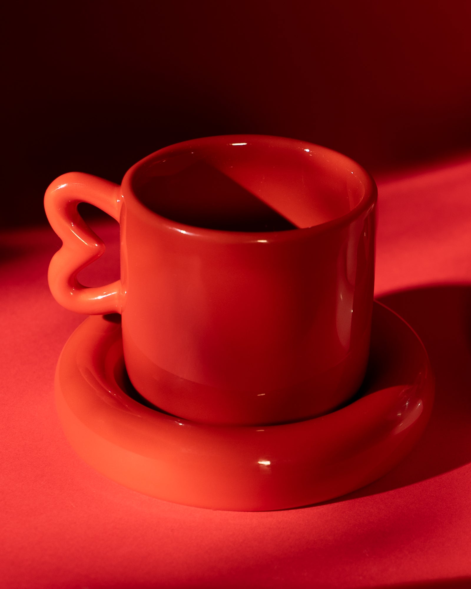 Rote Kaffeetasse mit herzförmigem Henkel auf rotem Untergrund, von einer Lichtquelle stilvoll beleuchtet.