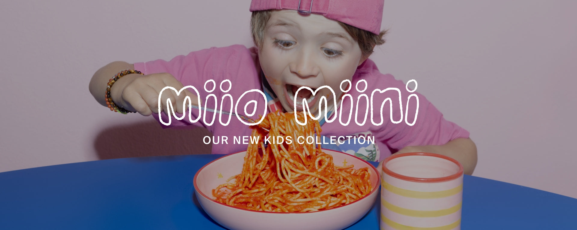 Kind isst begeistert Spaghetti, trägt rosa Kleidung und Mütze, mit buntem Becher daneben, Text: "Miio Miini Our New Kids Collection".