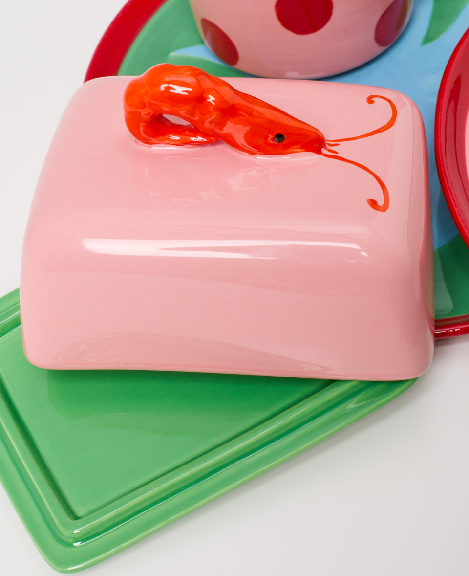 Eine rosa Butterdose mit einem roten Garnelengriff auf grünen und roten Keramiktellern.
