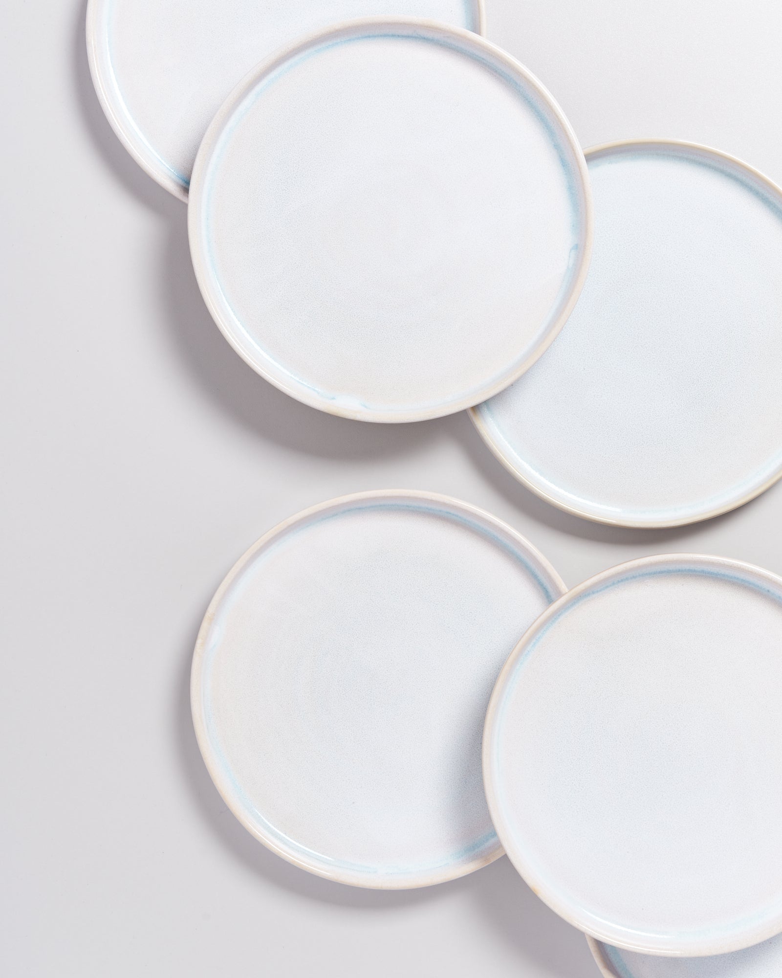 Sechs weiße Keramikteller mit leichtem blauen Rand liegen überlappend auf einer hellgrauen Fläche.