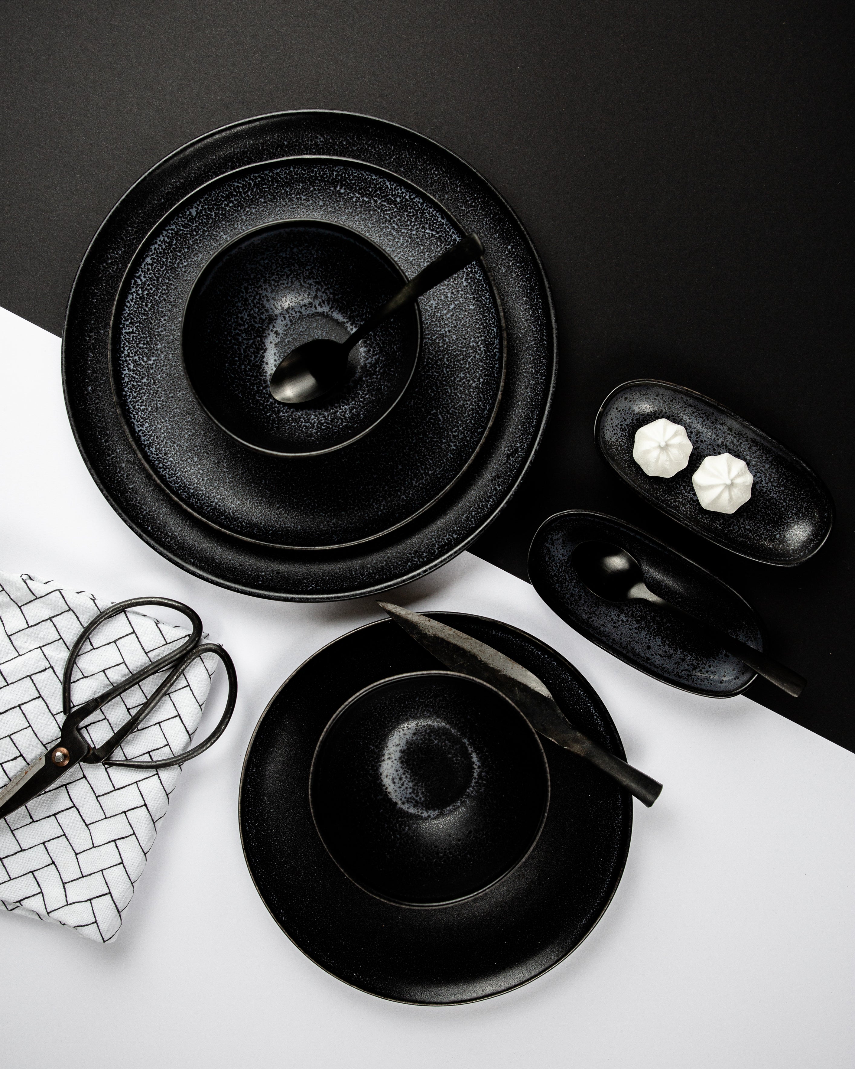 Schwarzes Geschirr mit Löffeln, auf schwarzem und weißem Hintergrund arrangiert, neben einer Schere und Stoffserviette.