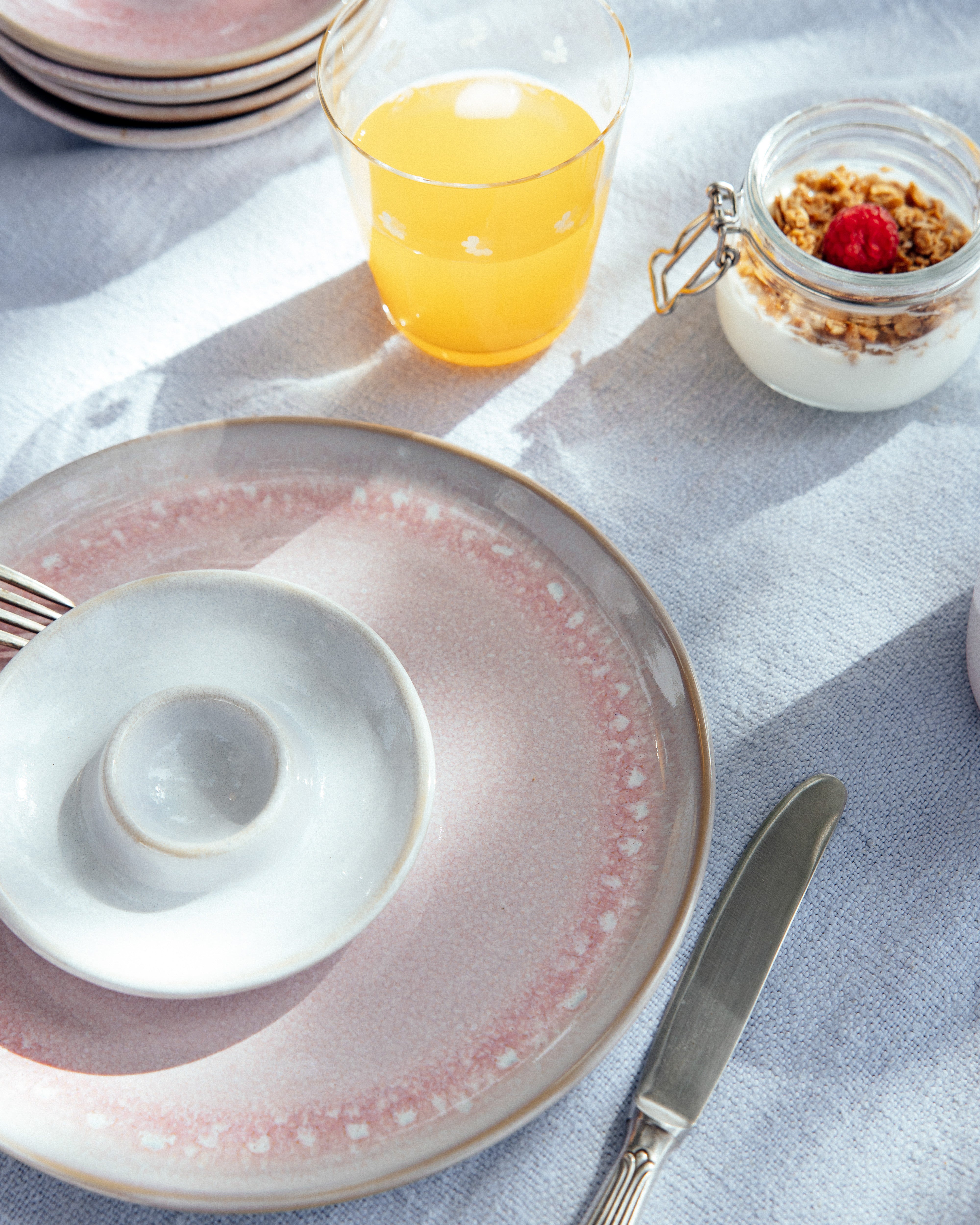 Frühstückstisch mit rosa Teller, kleinem weißen Schälchen, Glas Orangensaft und Joghurt mit Granola und Himbeere.