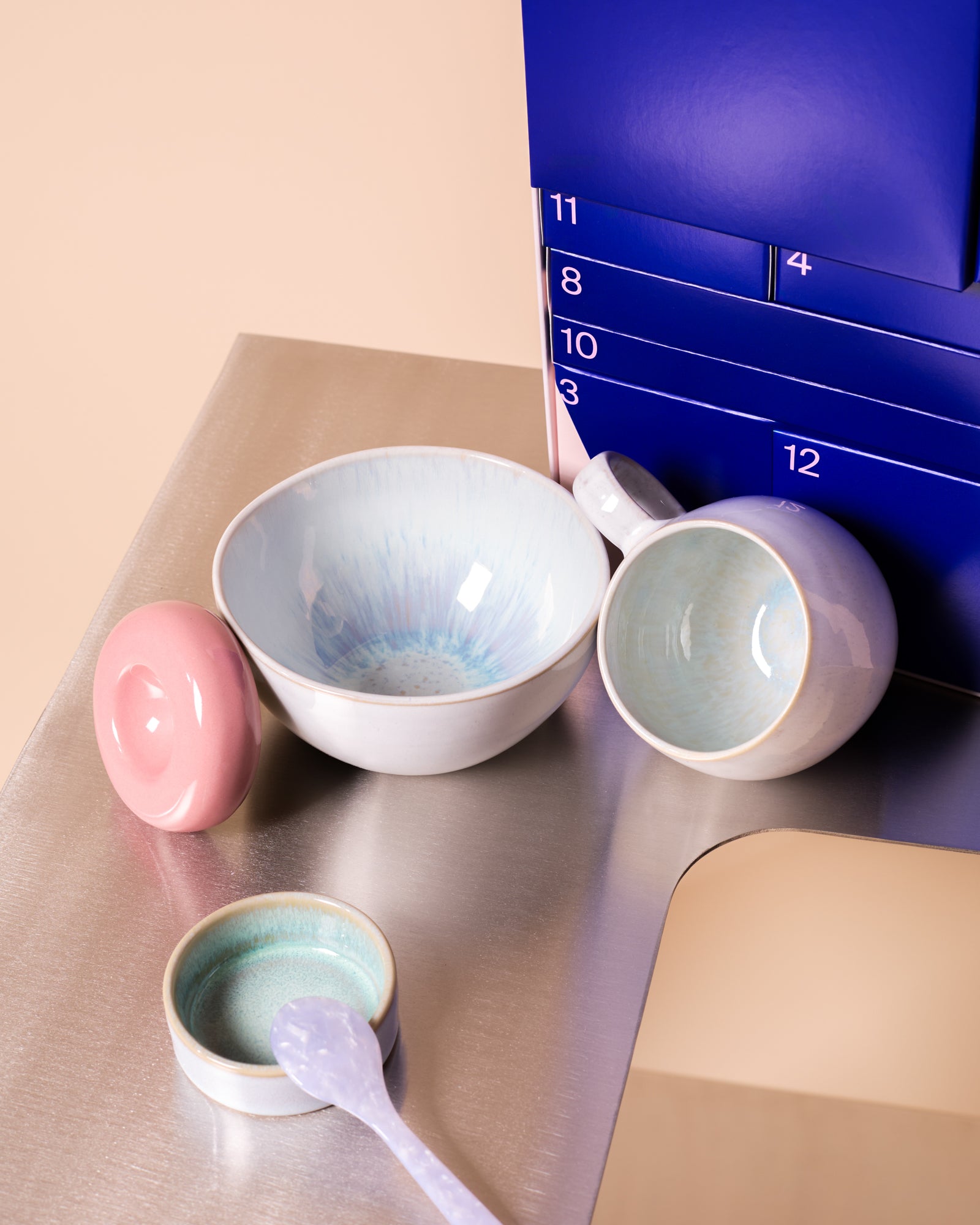 Keramikgeschirr mit pastellfarbenem Design auf einem Metalltisch, daneben ein blauer Kalender mit Zahlen.