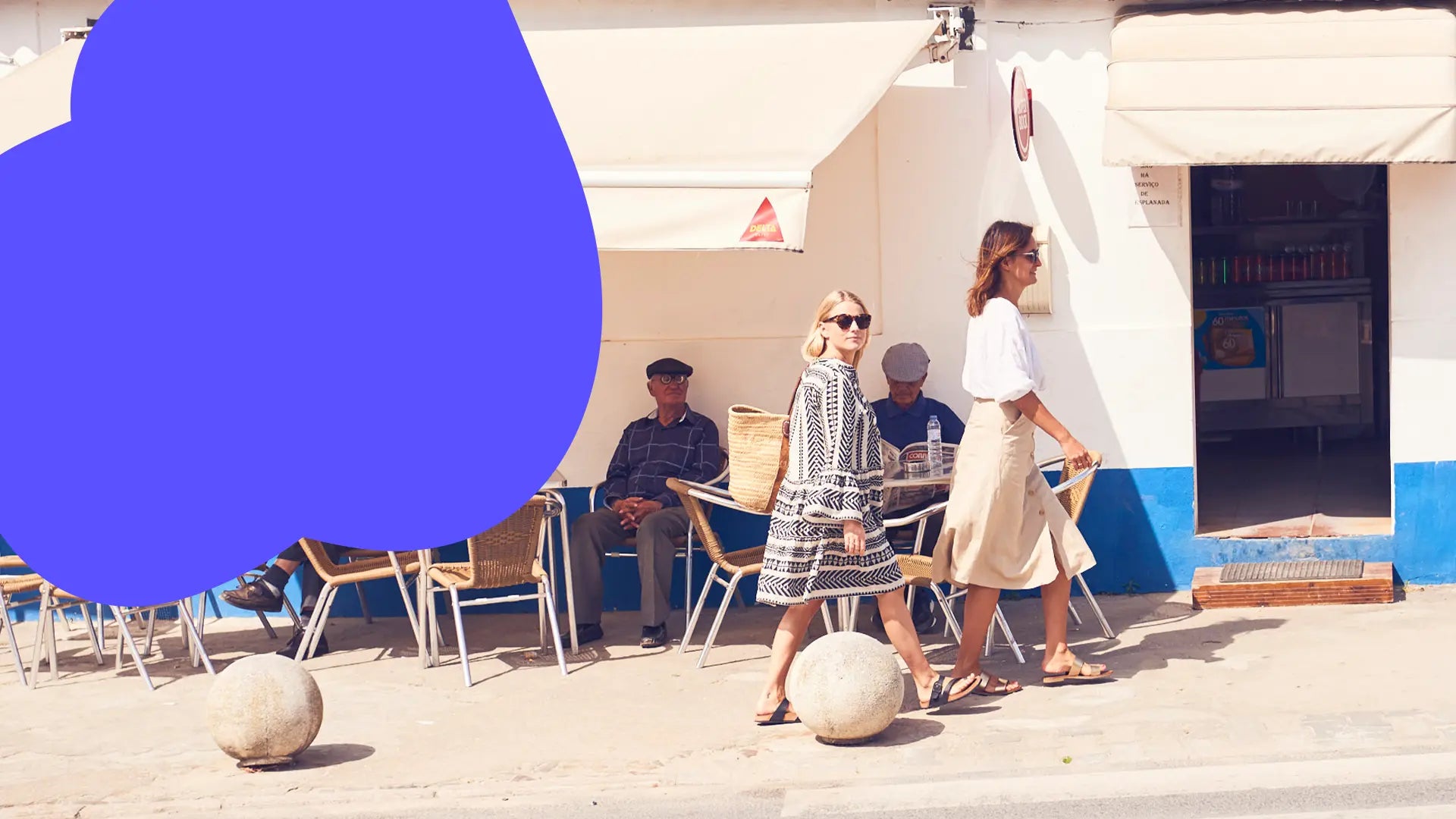 Zwei Frauen gehen an einem Café vorbei, in dem zwei ältere Männer draußen sitzen. Eine große blaue Form verdeckt Teil der Szene.