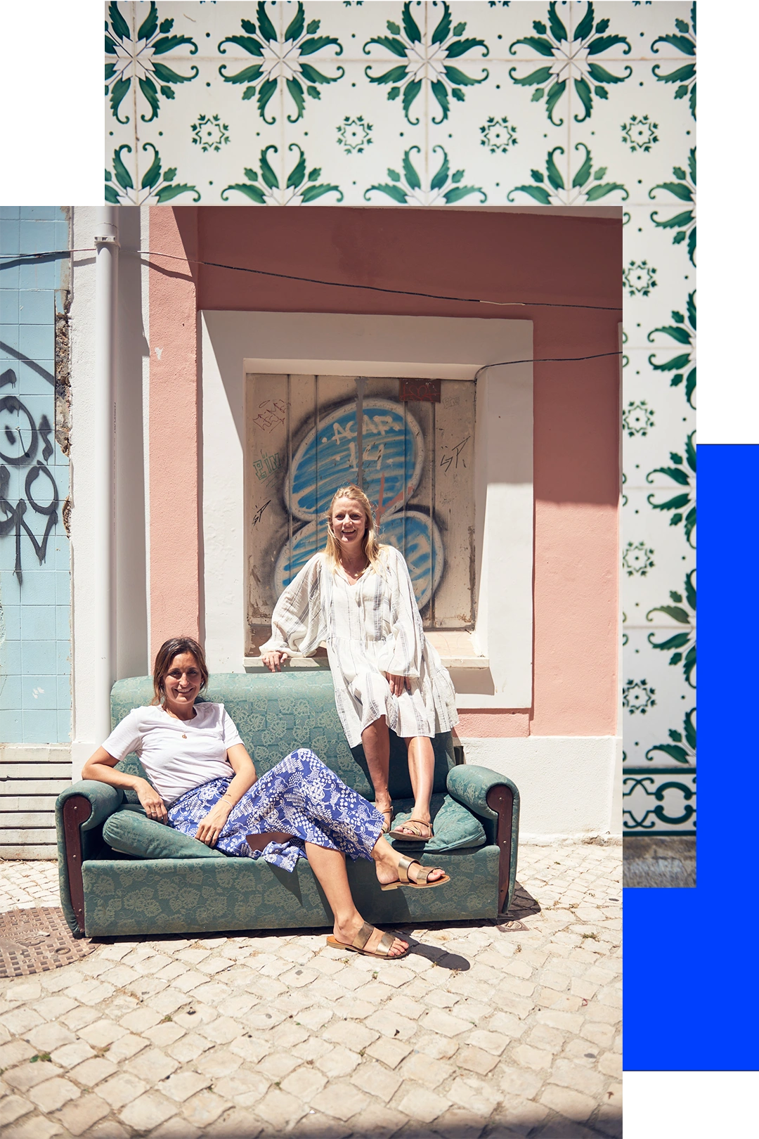 Zwei Frauen sitzen auf einem grünen Sofa vor einer rosa Wand mit Graffiti und Fliesenmustern im Hintergrund.