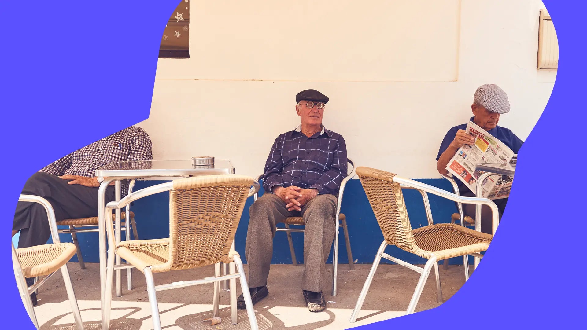 Drei ältere Männer sitzen auf einer Terrasse; einer liest eine Zeitung, die anderen sitzen mit gefalteten Händen.