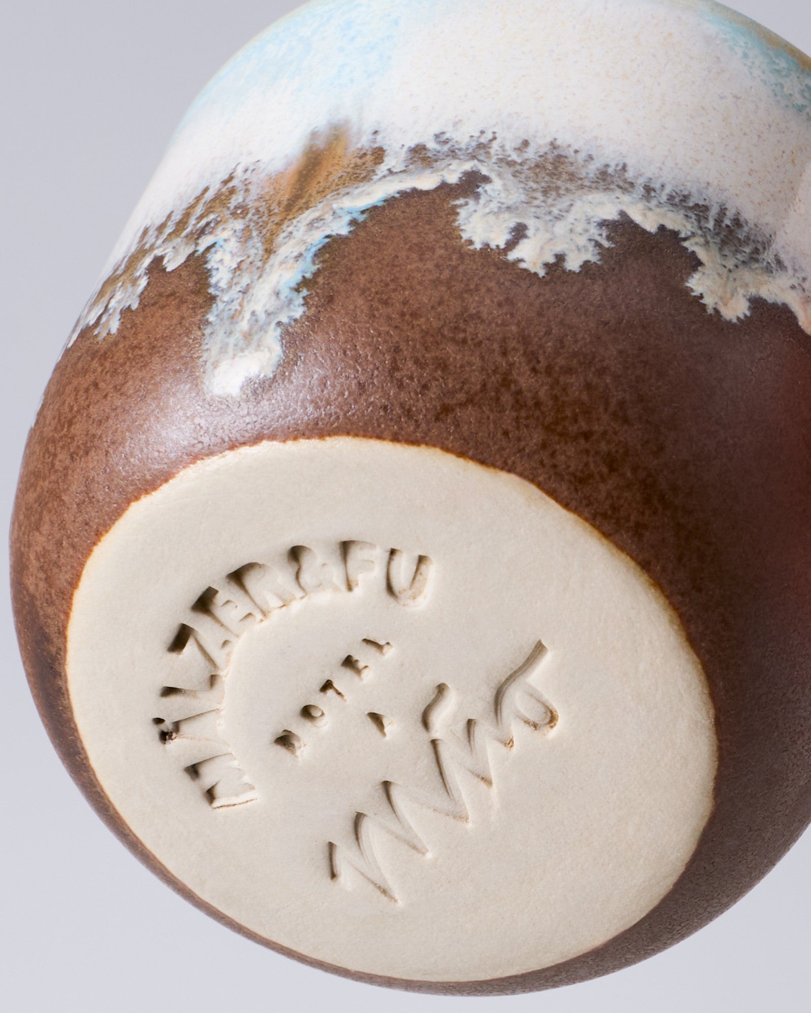 Detailansicht eines keramischen Bechers mit brauner und weißer Glasur sowie eingraviertem Schriftzug am Boden.