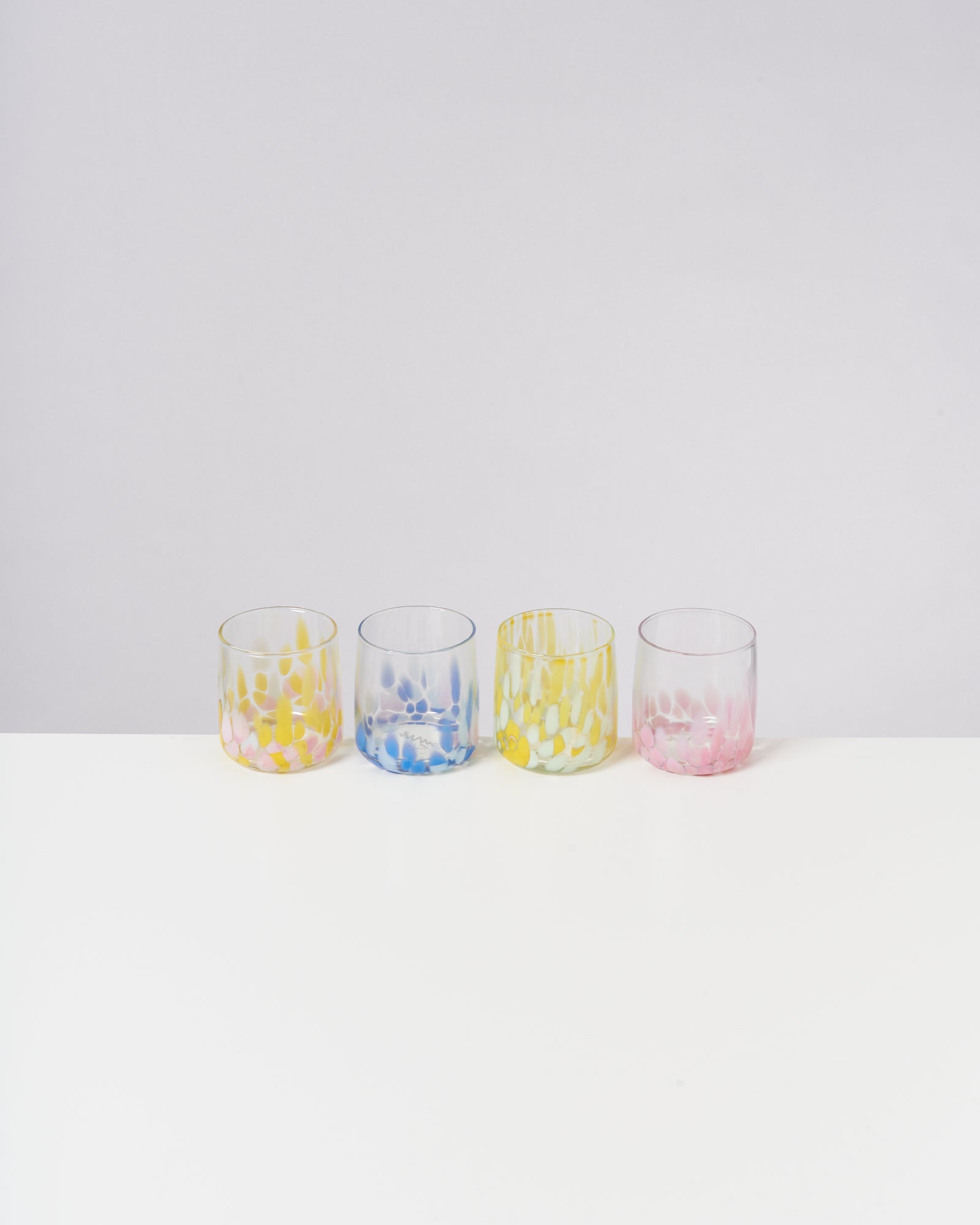 Vier bunte Gläser mit gelben, blauen und rosa Mustern stehen auf einem weißen Tisch vor hellem Hintergrund.