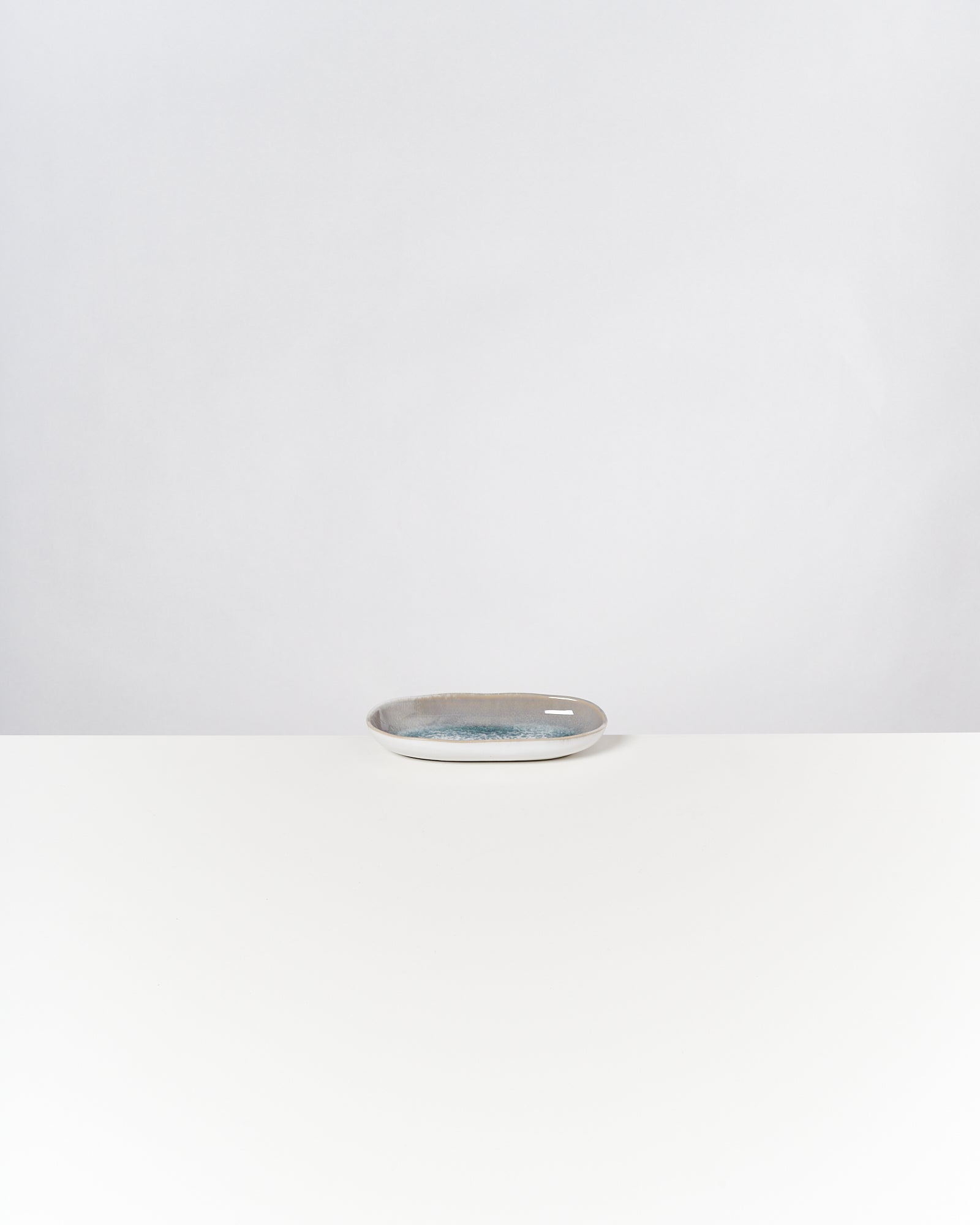 Ein kleiner ovaler Teller mit graublauer Glasur steht auf einem weißen Tisch vor einem grauen Hintergrund.