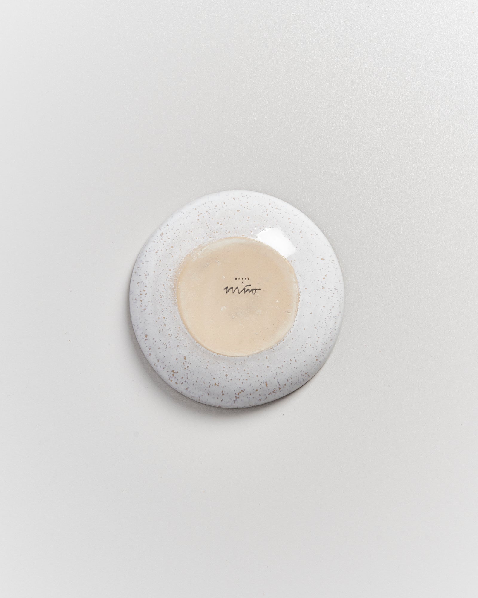 Die Unterseite eines weißen, gesprenkelten Keramiktellers mit einer mittigen Signatur auf heller Oberfläche.