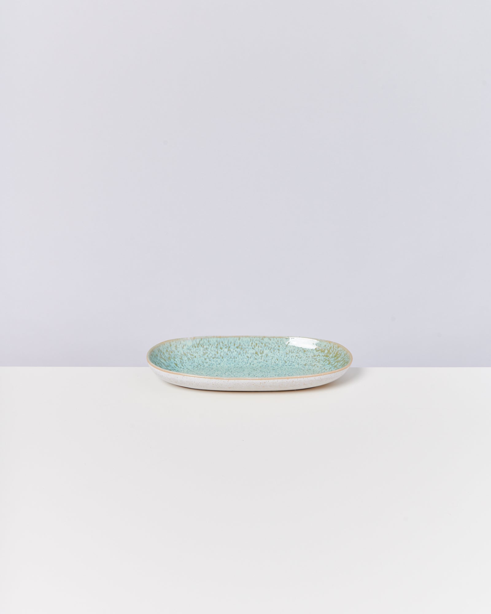 Ein flacher, ovaler, türkisfarbener Keramikteller steht auf einer weißen Oberfläche vor einem grauen Hintergrund.
