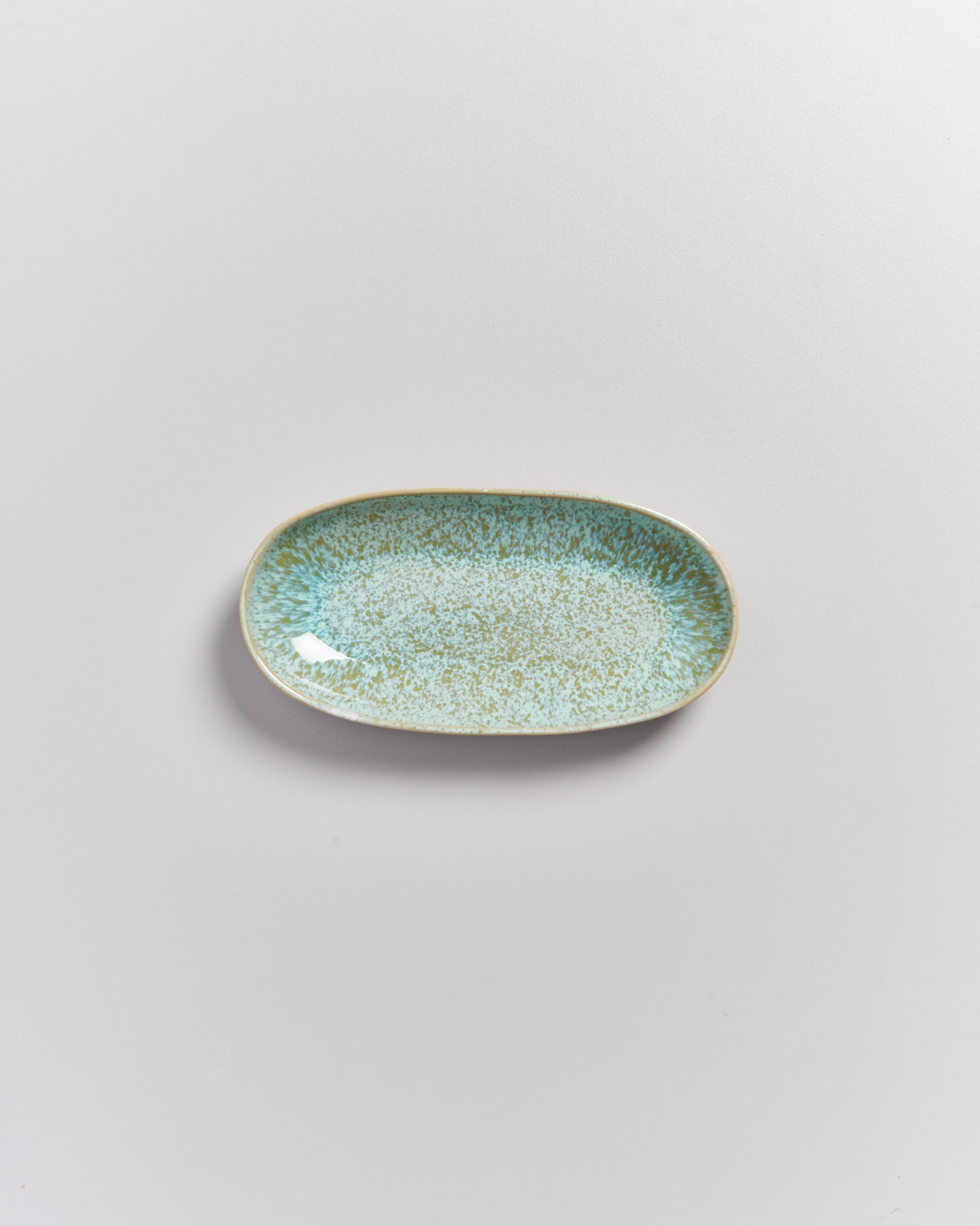 Ovale Keramikplatte mit blau-grüner Glasur und gesprenkeltem Muster auf hellem Untergrund.