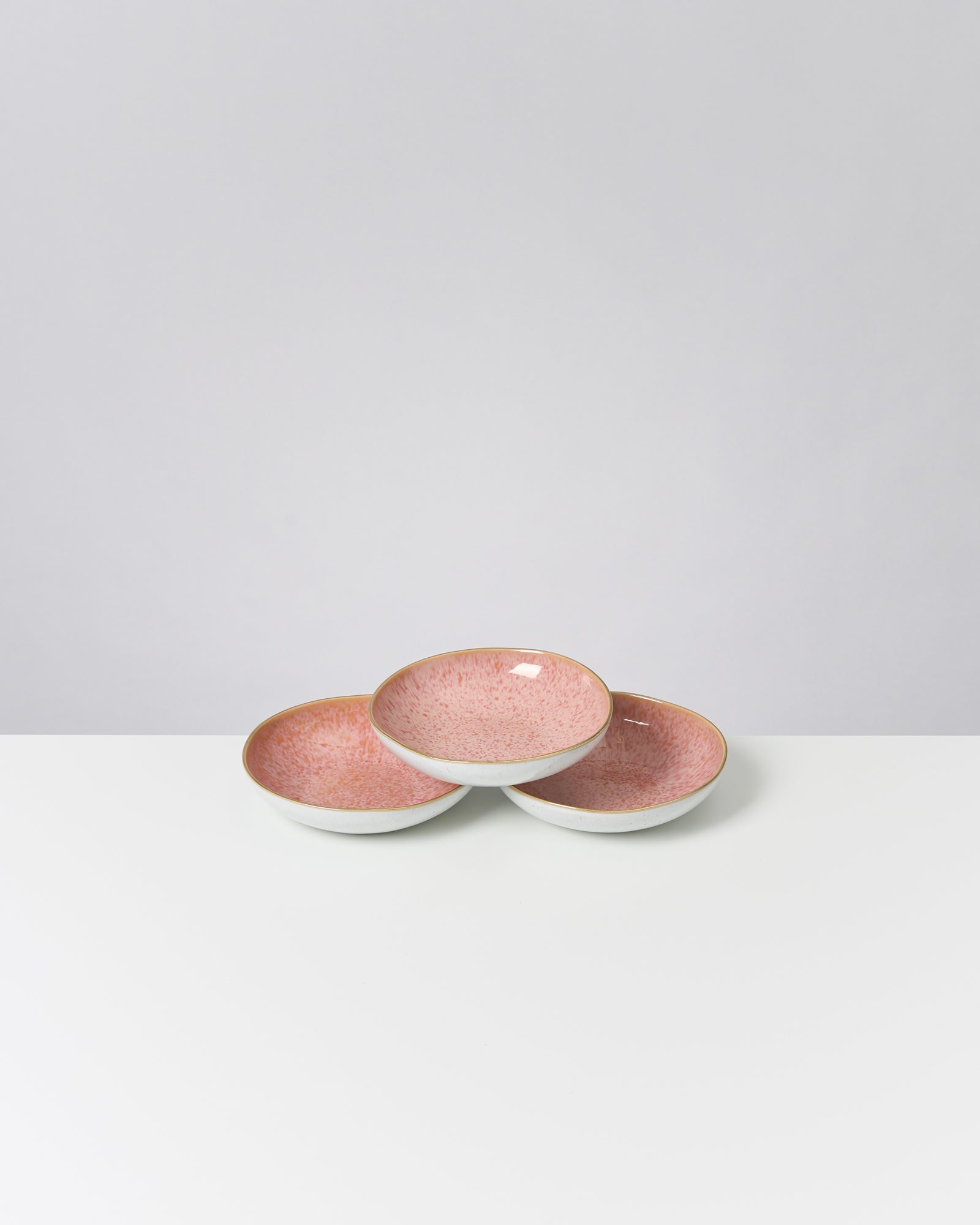 Drei rosafarbene Schalen aus Keramik mit gesprenkeltem Muster stehen auf einem weißen Tisch vor grauem Hintergrund.
