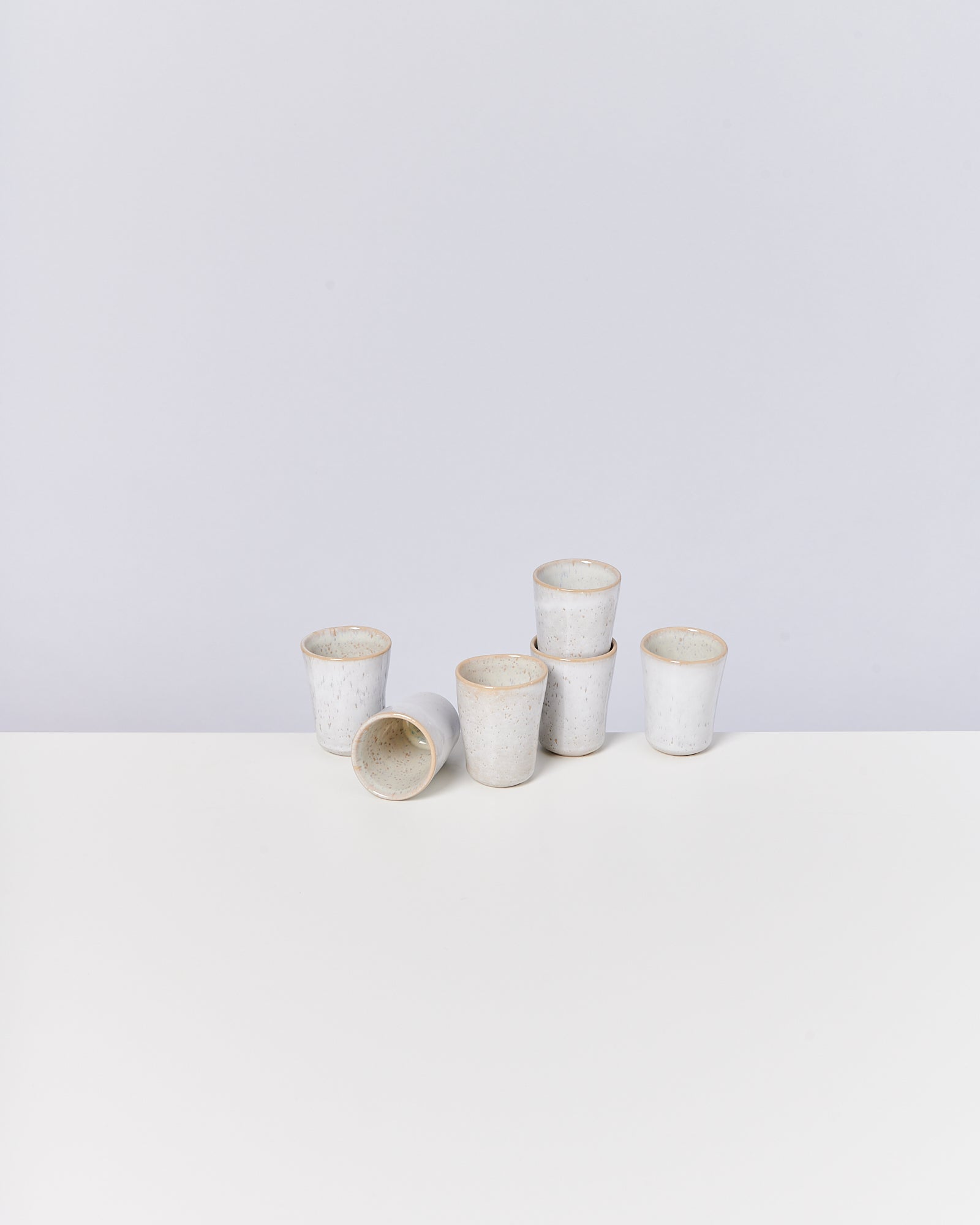 Sechs weiße Keramikbecher, einer liegend, auf einer weißen Oberfläche vor grauem Hintergrund.