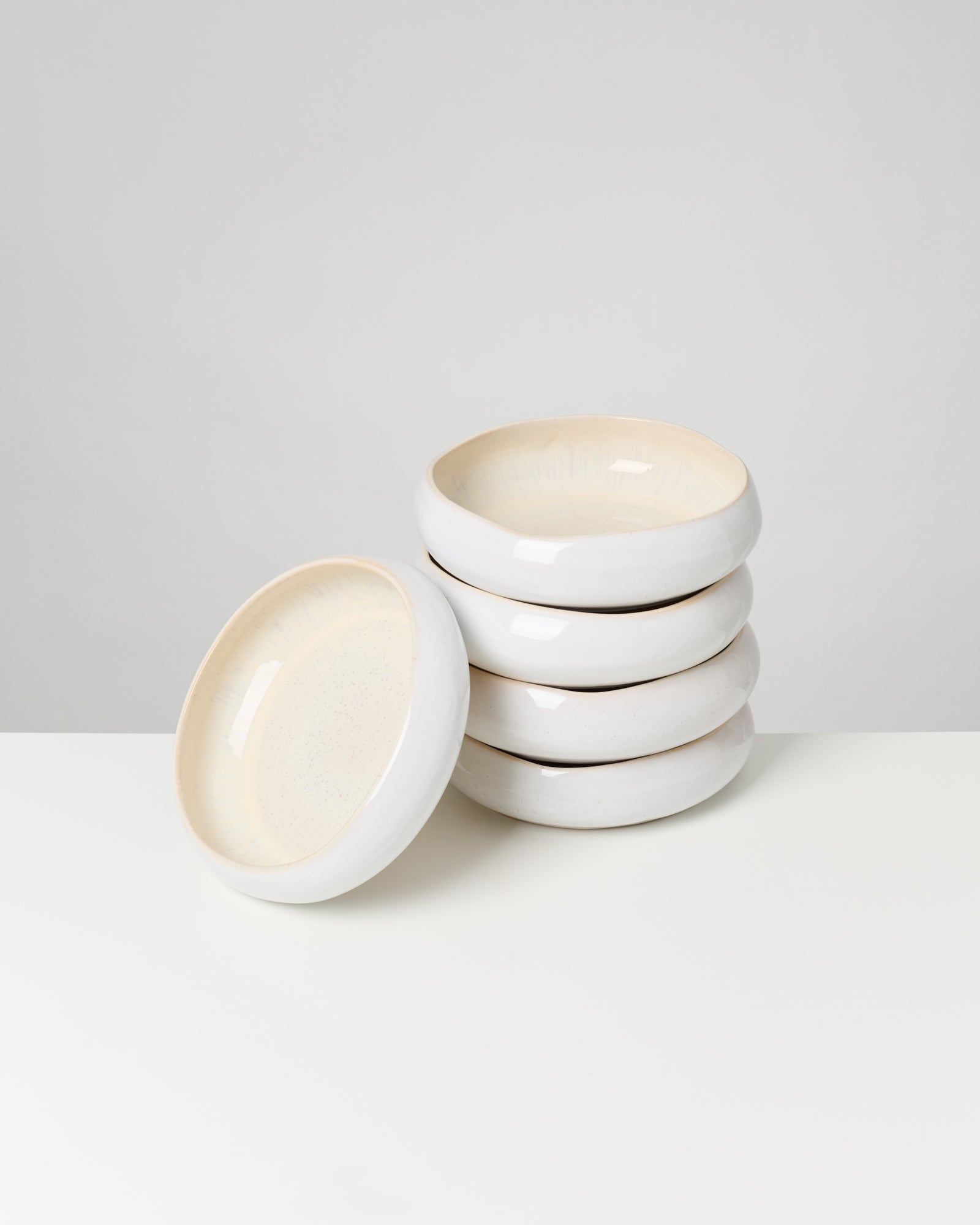 Ein Stapel von sechs weißen, runden Keramiktellern, einer lehnt an der Seite, auf einem weißen Tisch vor grauem Hintergrund.