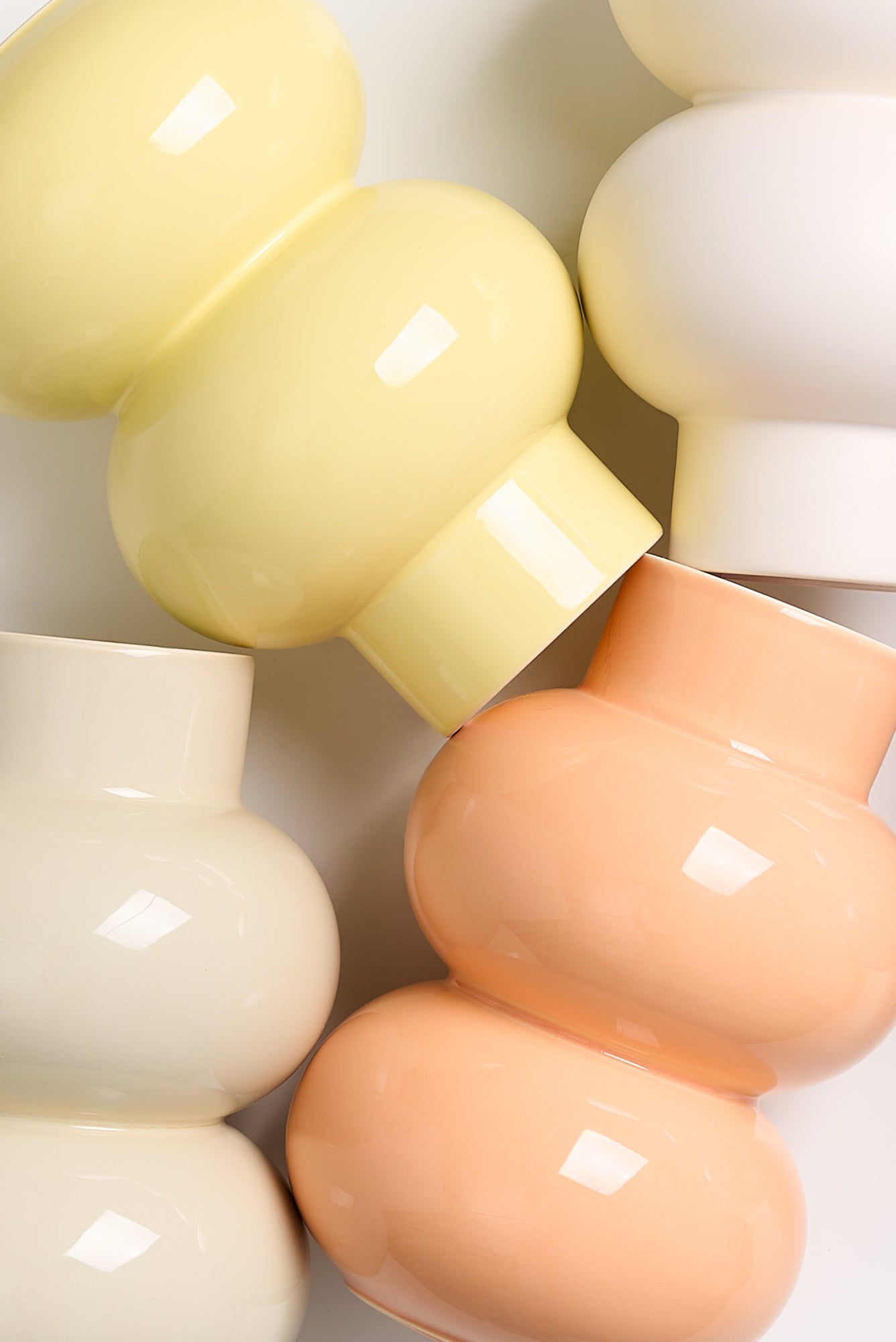 Vier verschiedenfarbige, glänzende Vasen in Pastelltönen liegen ineinander verschlungen auf einer weißen Oberfläche.