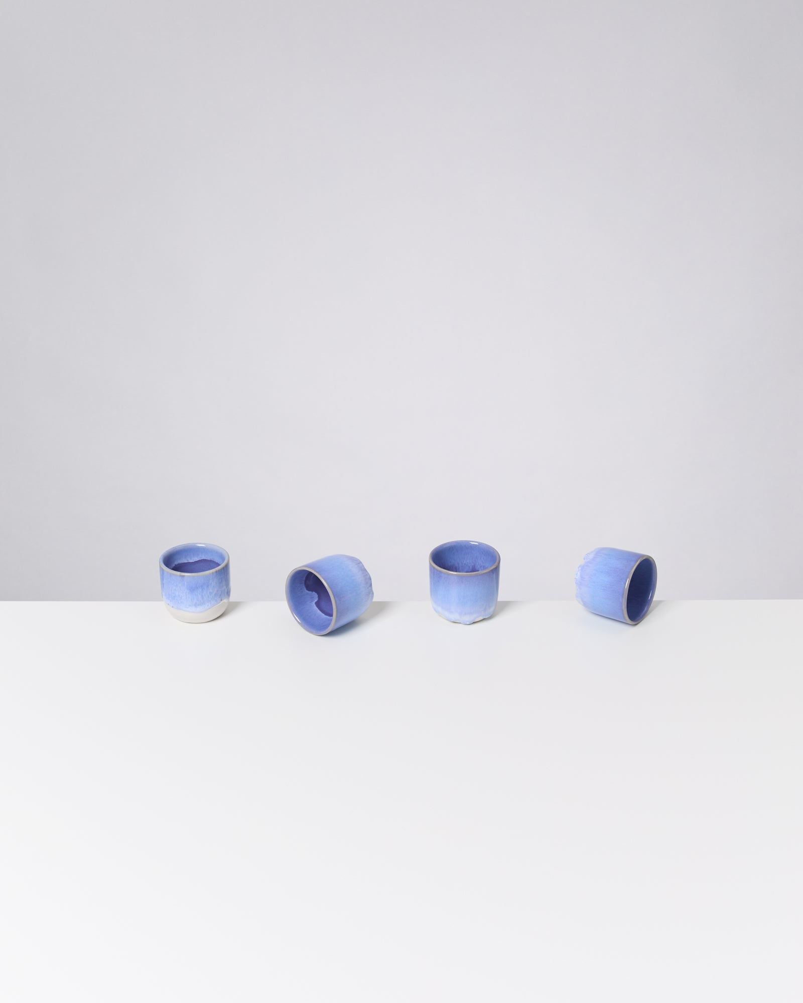 Vier blaue Keramiktassen stehen in einer Reihe auf einer weißen Oberfläche vor einem hellgrauen Hintergrund.