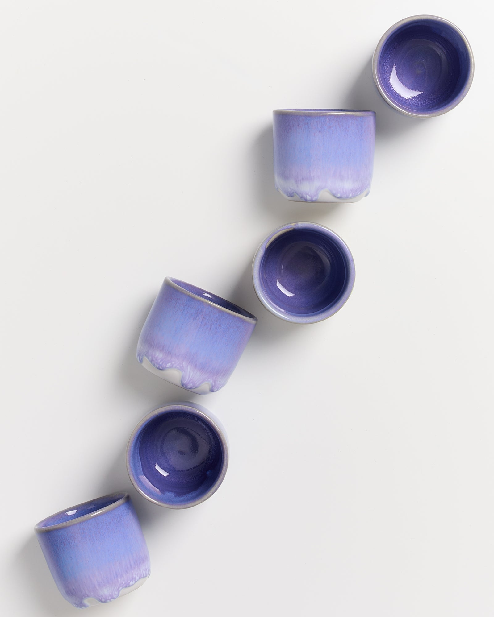 Sechs violette Tassen in einer schrägen Linie auf weißem Hintergrund angeordnet.