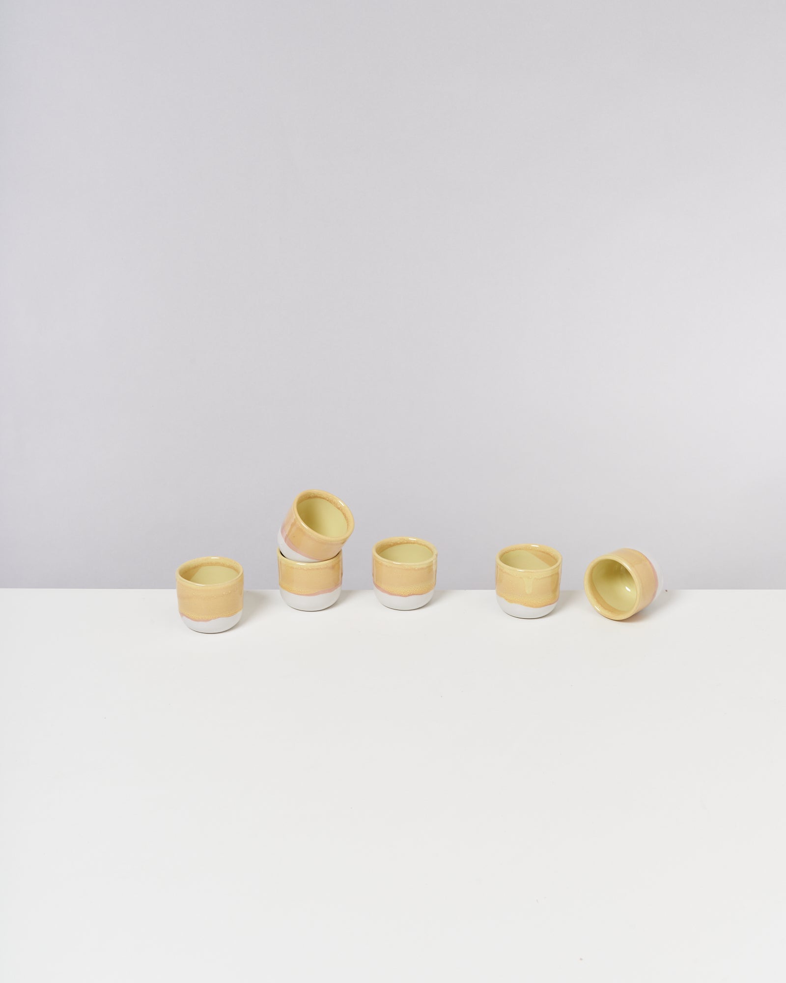 Sechs gelb-weiße Keramikbecher sind auf einer weißen Oberfläche angeordnet, einige liegen seitlich.