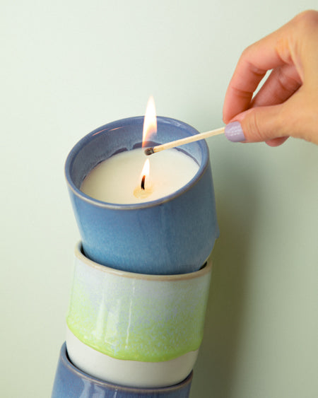Hand entzündet mit einem Streichholz eine Kerze in einem mehrfarbigen Keramikgefäß auf einem mintgrünen Hintergrund.