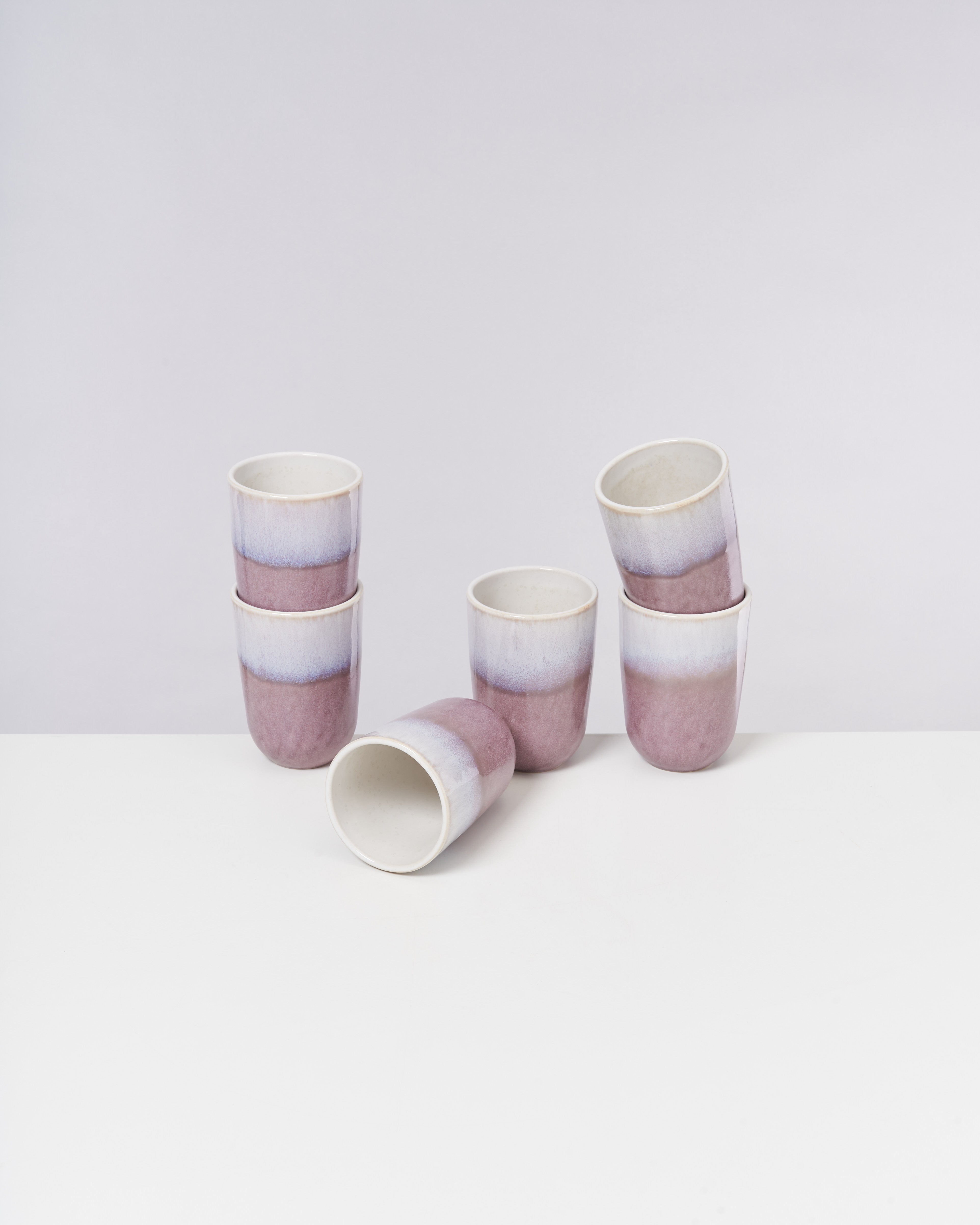 Sechs lila-weiße Keramikbecher, fünf aufrecht und einer liegend, auf einem weißen Tisch vor einem grauen Hintergrund.
