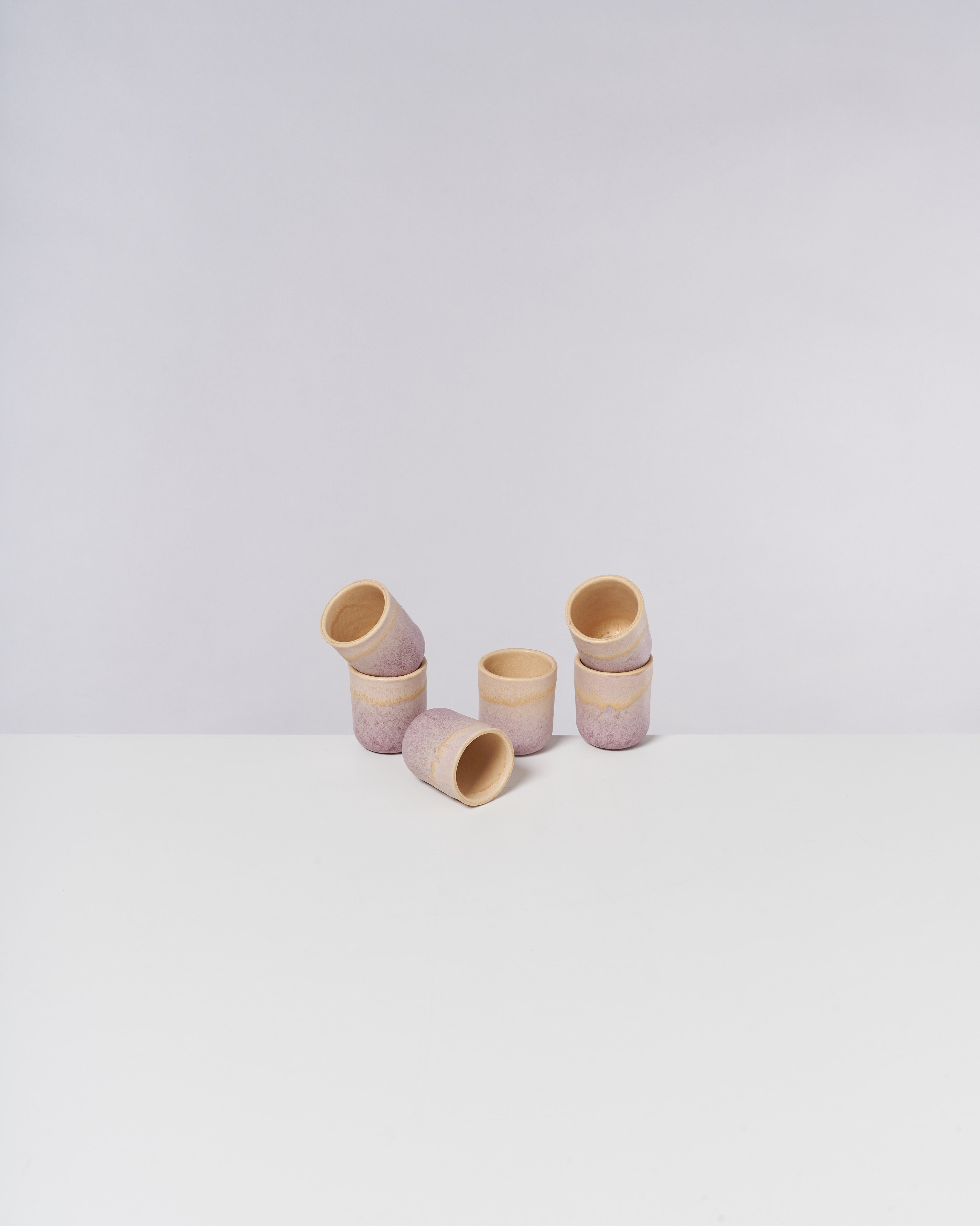 Sieben stapelbare Becher aus Ton mit einem zarten rosa Muster stehen auf einer weißen Oberfläche vor grauem Hintergrund.