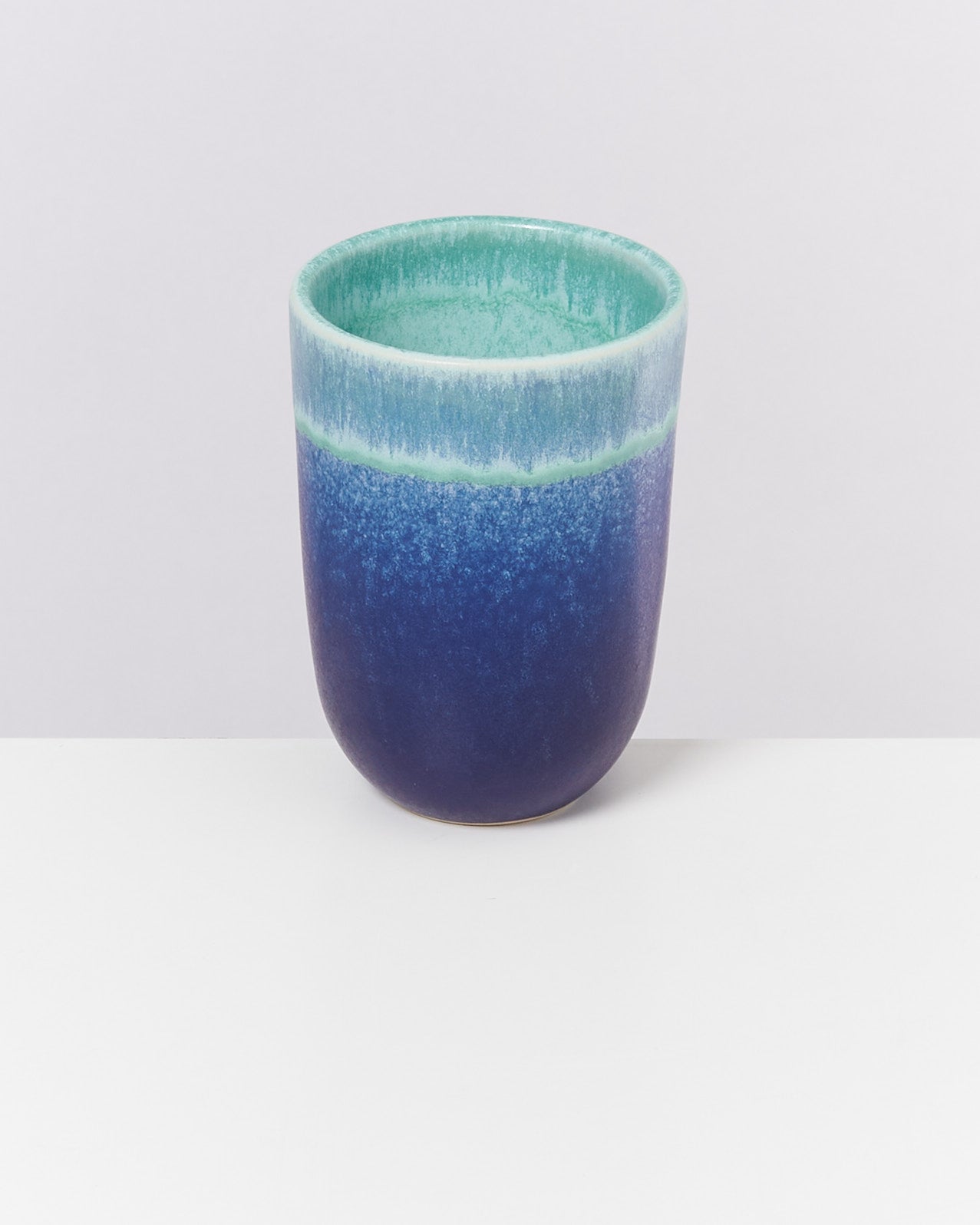Blauer und grüner Keramikbecher auf einer weißen Oberfläche vor einem hellgrauen Hintergrund.
