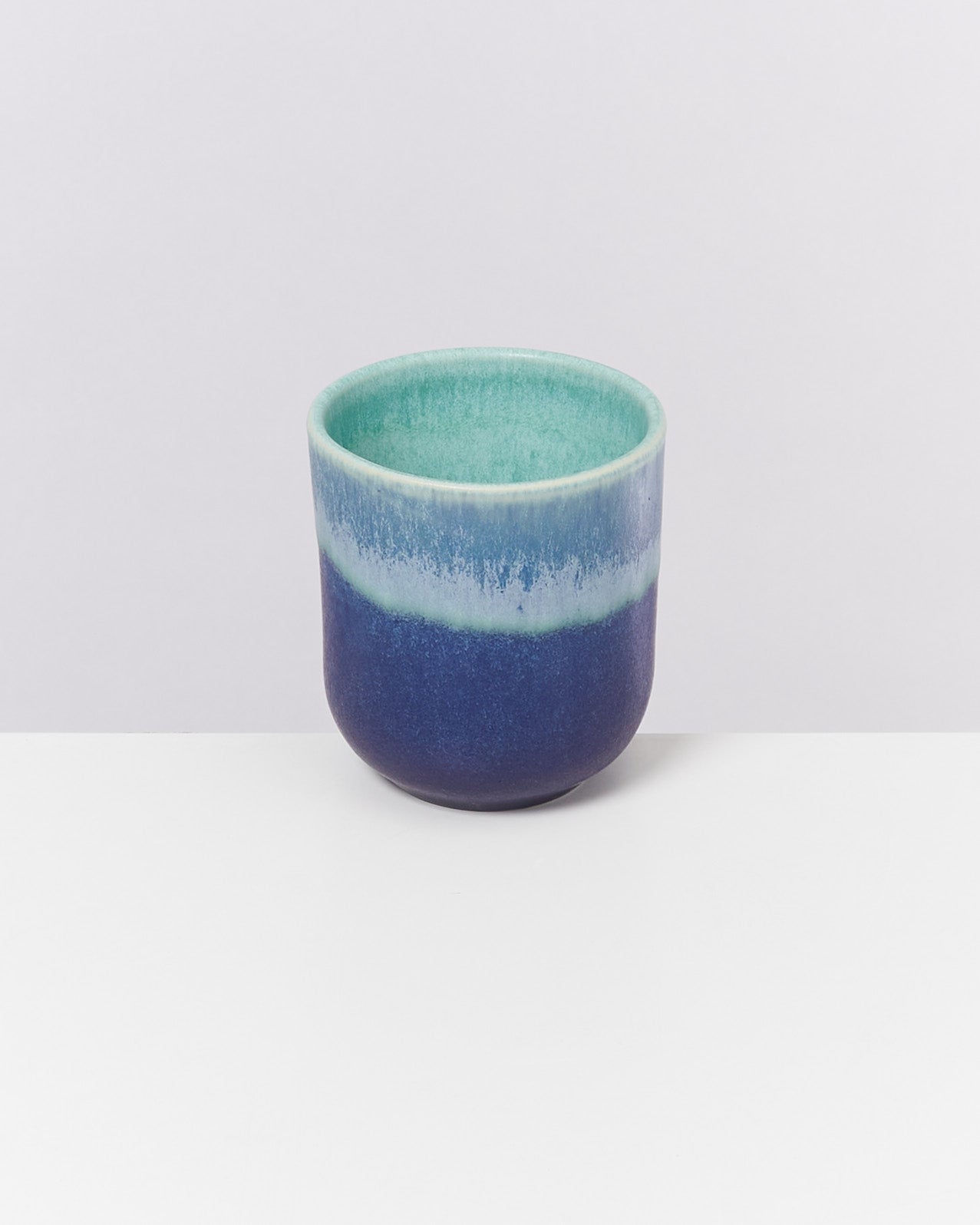 Kleine Keramikvase in Blautönen, stehend auf einem weißen Tisch vor einem hellen Hintergrund.