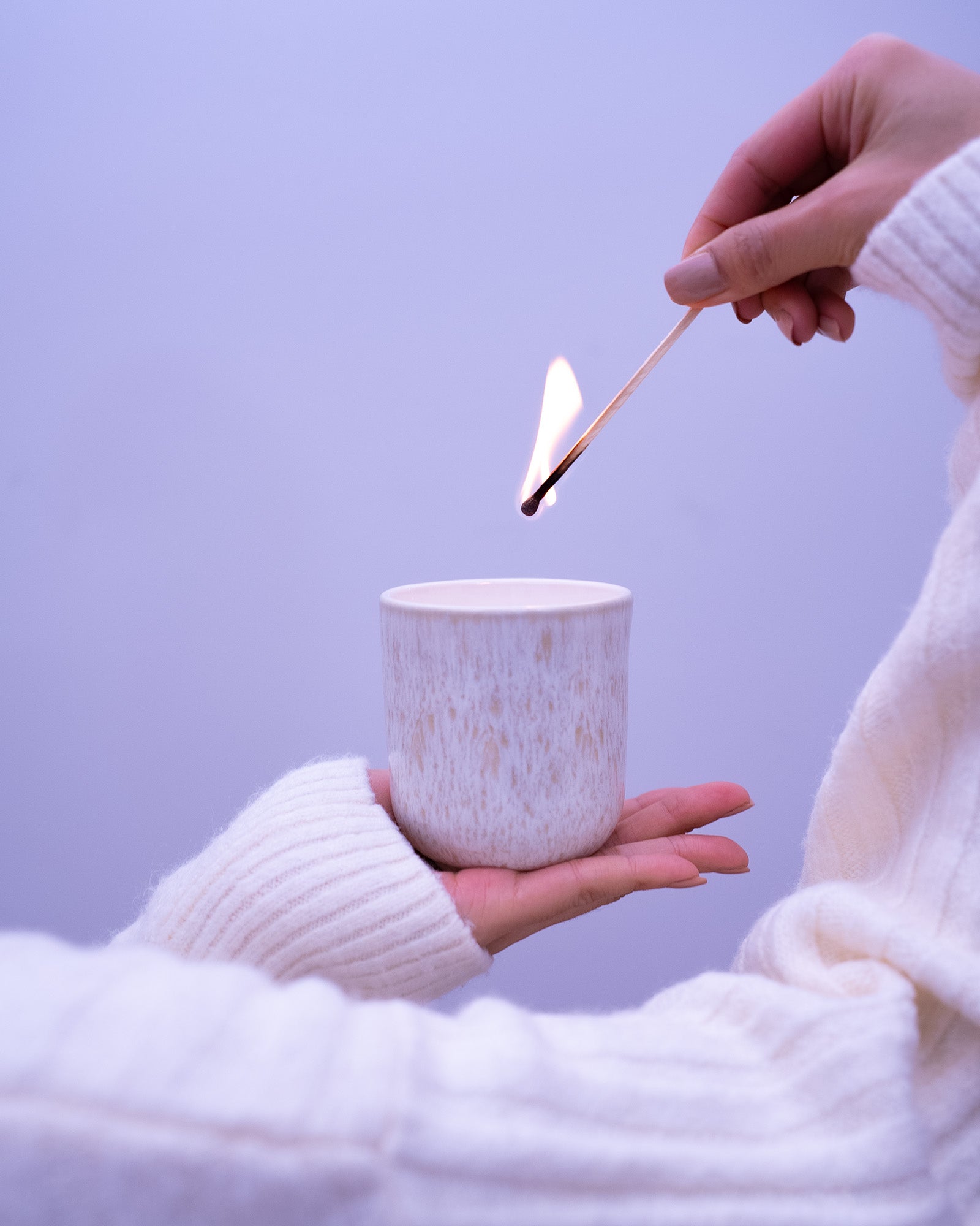 Eine Person hält eine Kerze in der Hand und zündet sie mit einem brennenden Streichholz an, vor einem lilafarbenen Hintergrund.