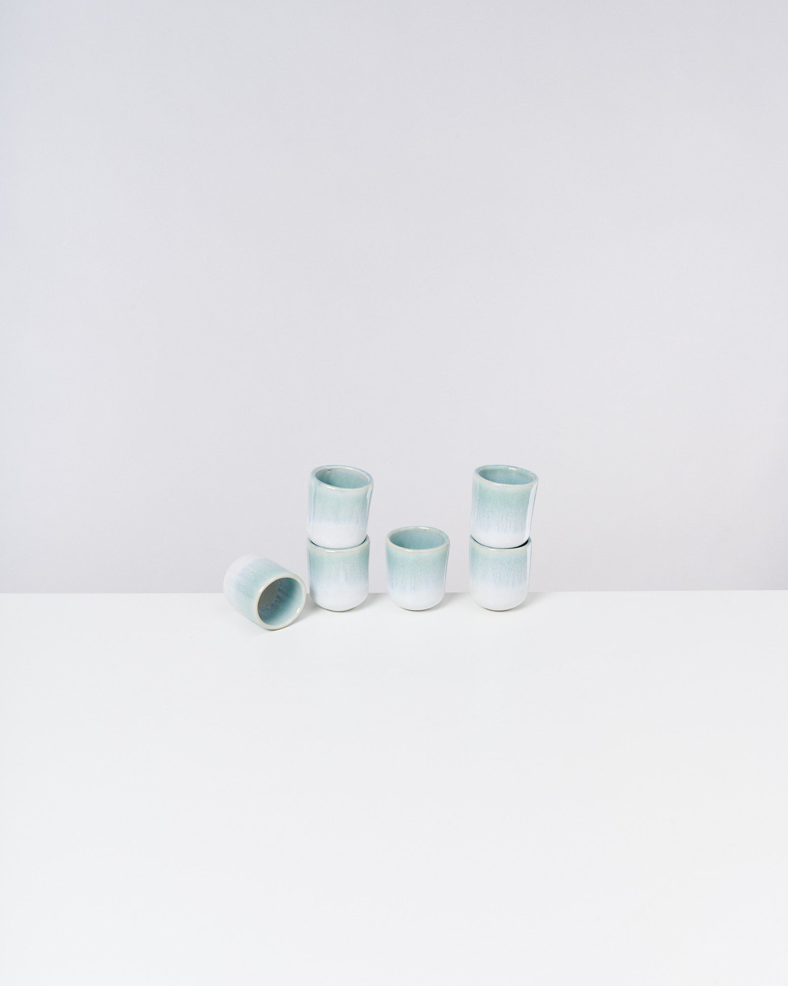 Sechs hellblaue Keramikbecher stehen und ein Becher liegt auf einer weißen Oberfläche vor einem weißen Hintergrund.