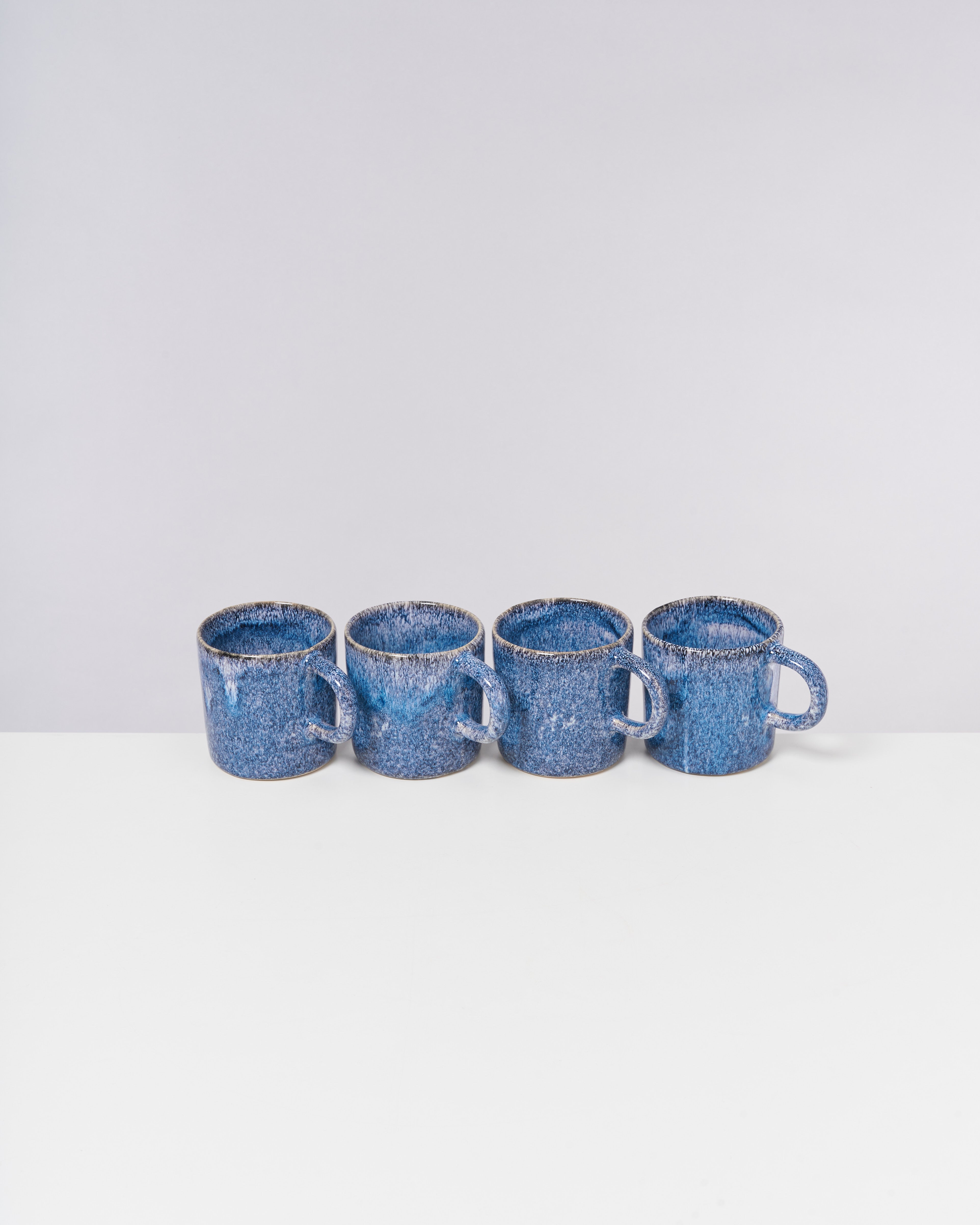 Vier blaue Keramiktassen stehen in einer Reihe auf einem weißen Tisch vor einem grauen Hintergrund.