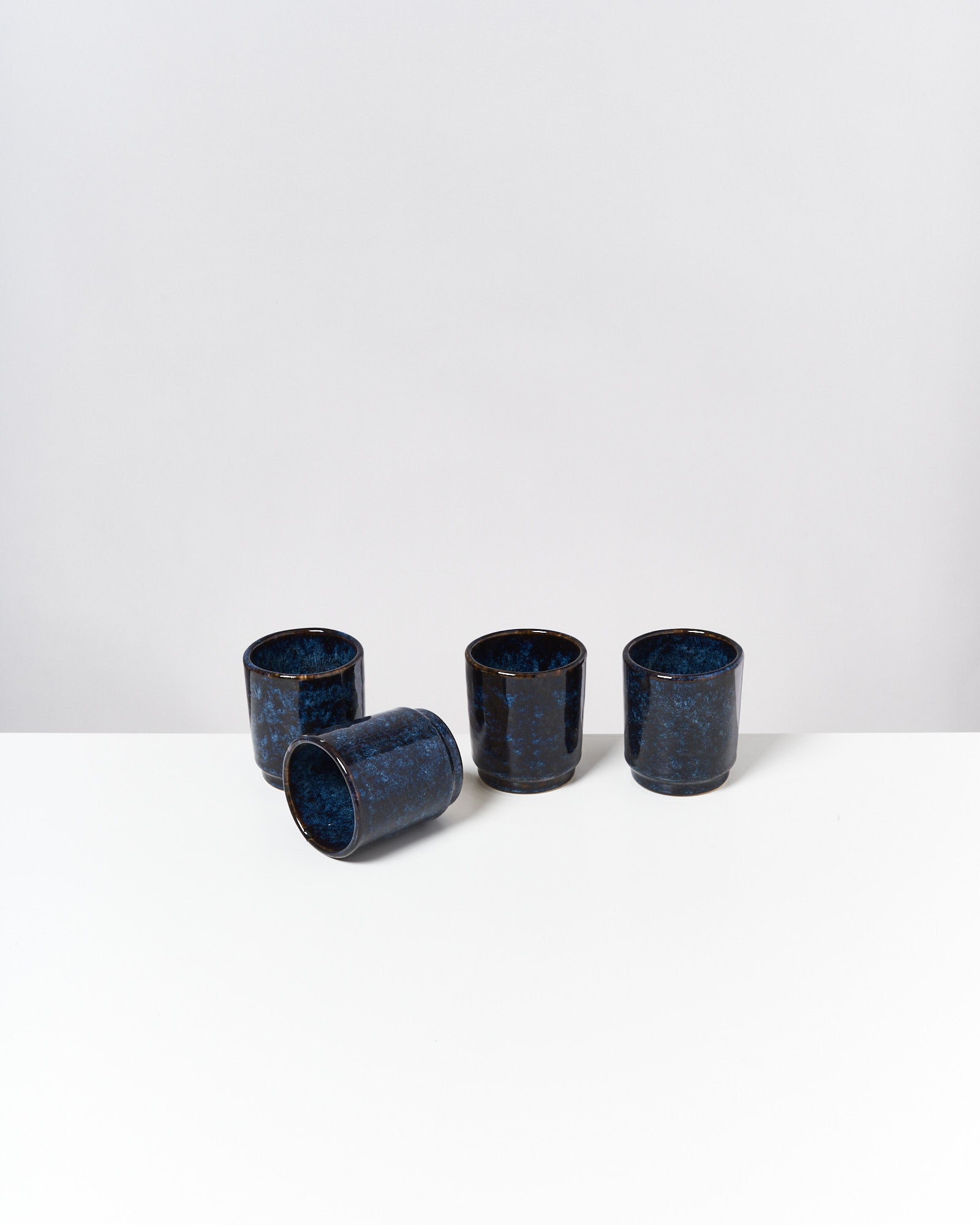 Vier blaue Keramikbecher, einer liegend, auf einer weißen Oberfläche gegen einen grauen Hintergrund.