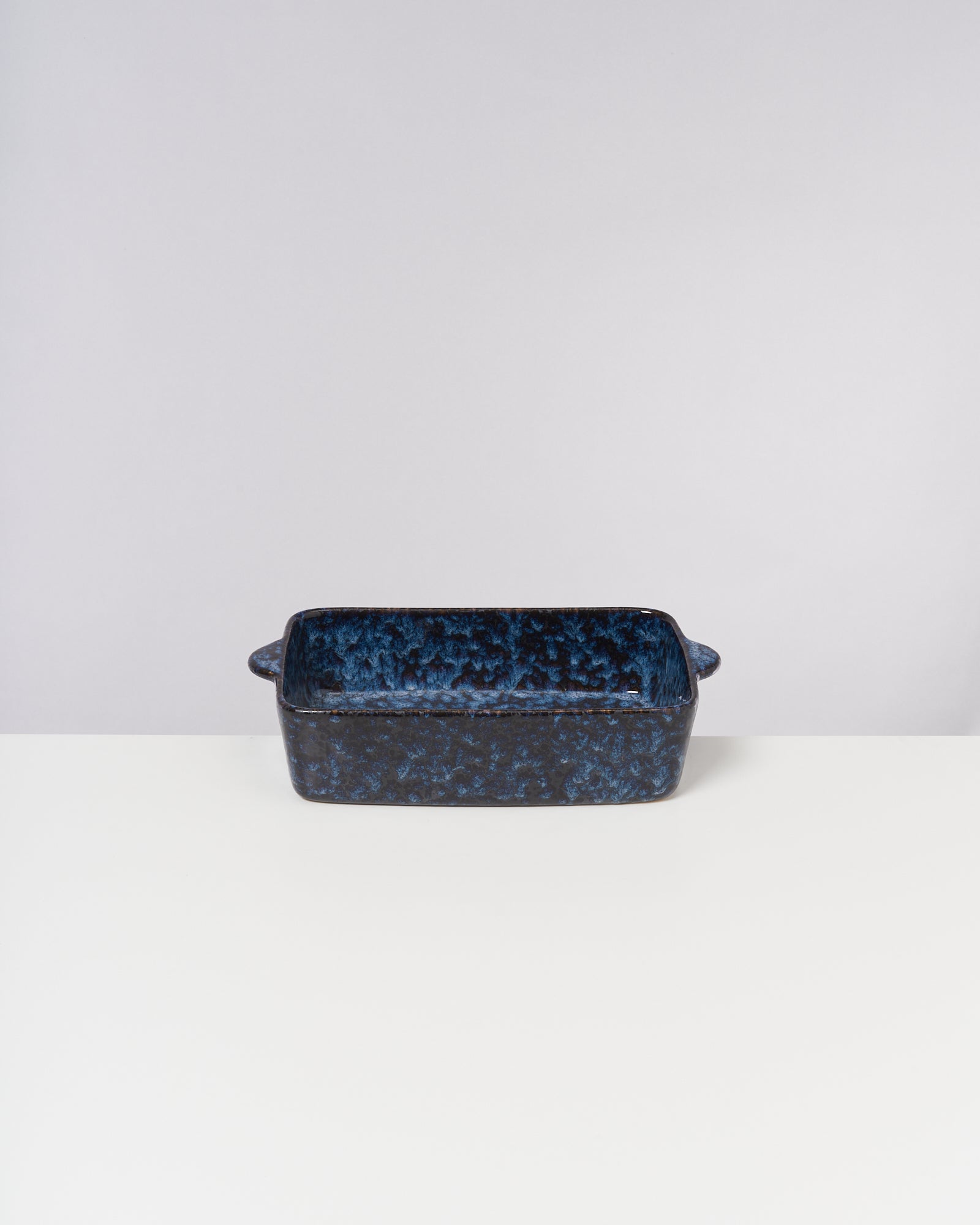 Blaue Keramikauflaufform mit Henkeln steht auf einem weißen Tisch vor neutralem Hintergrund.