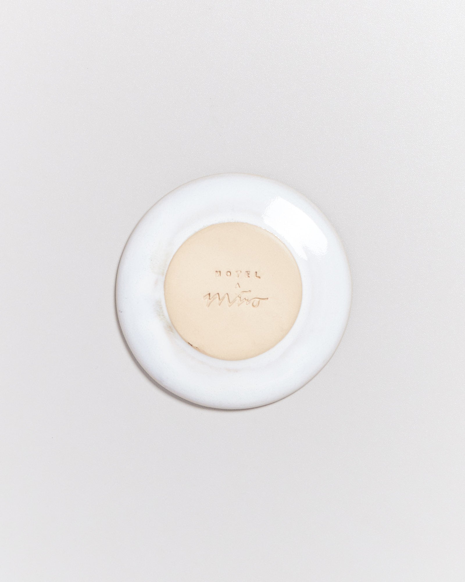 Kleine weiße Untertasse mit der Aufschrift "Hotel a Miro" in der Mitte auf hellem Hintergrund.