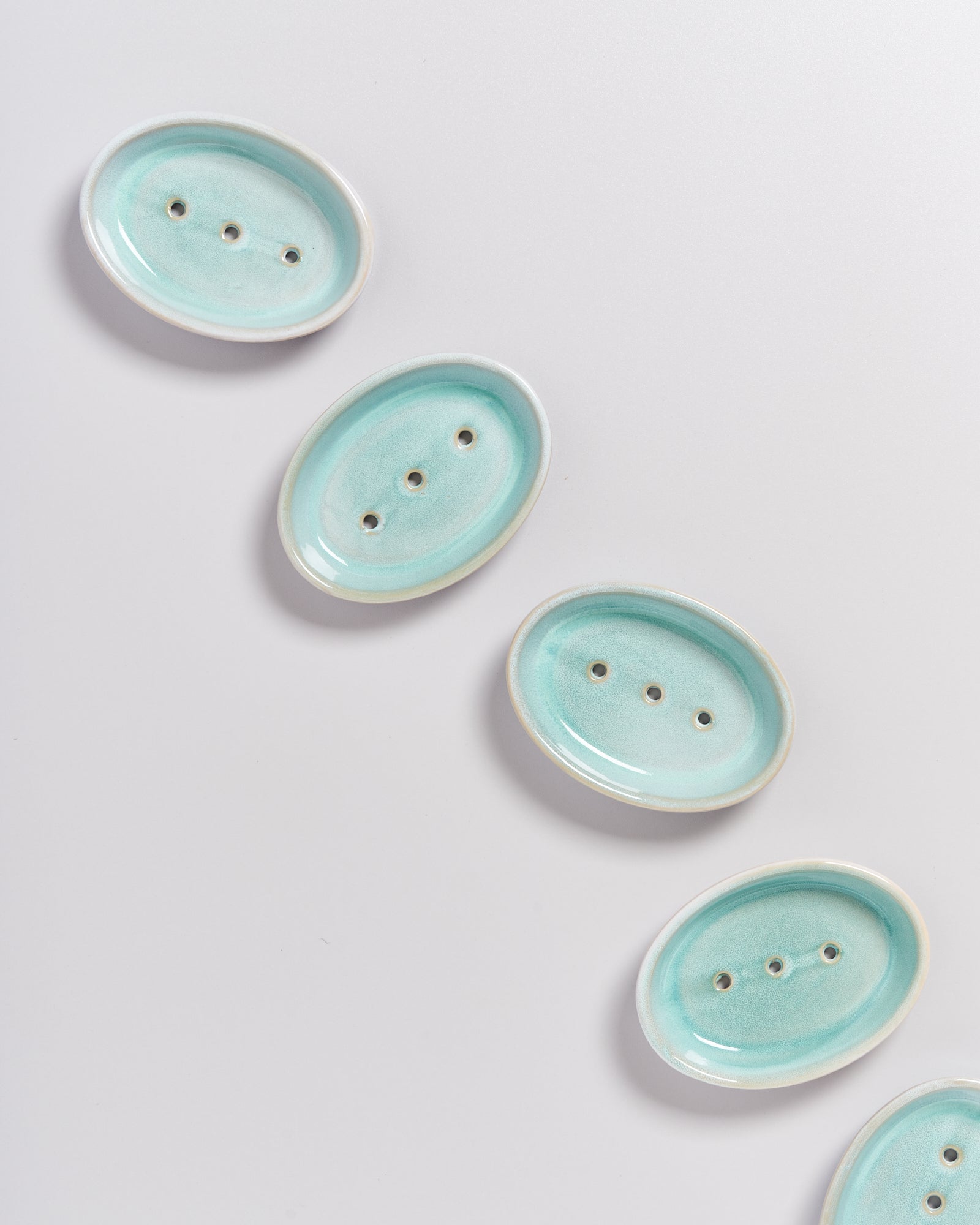 Fünf ovale, hellblaue Seifenschalen mit drei Löchern, diagonal auf einem weißen Hintergrund angeordnet.