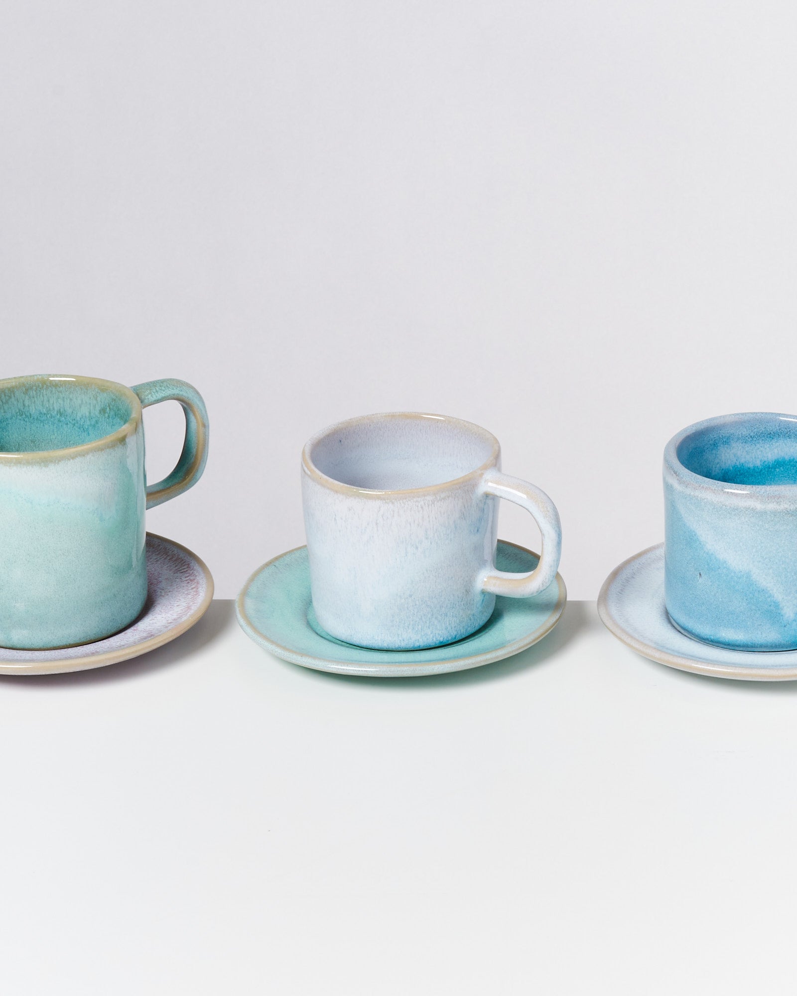 Drei pastellfarbene, handgefertigte Tassen auf Untertassen in einem schlichten, modernen Stil auf weißem Hintergrund.