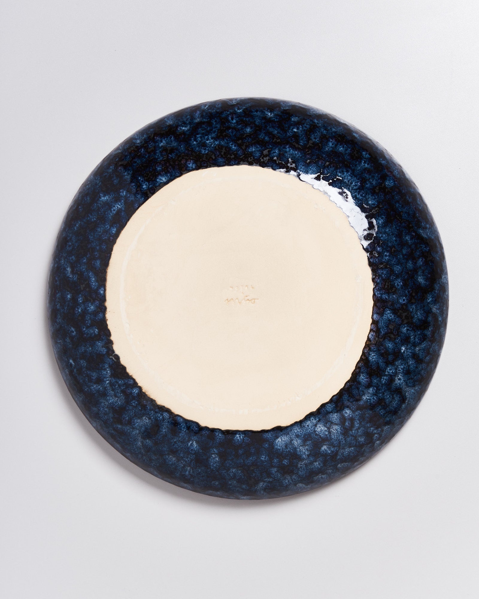 Unterseite eines runden Tellers mit dunkelblauem Rand und heller Mitte auf weißem Hintergrund.